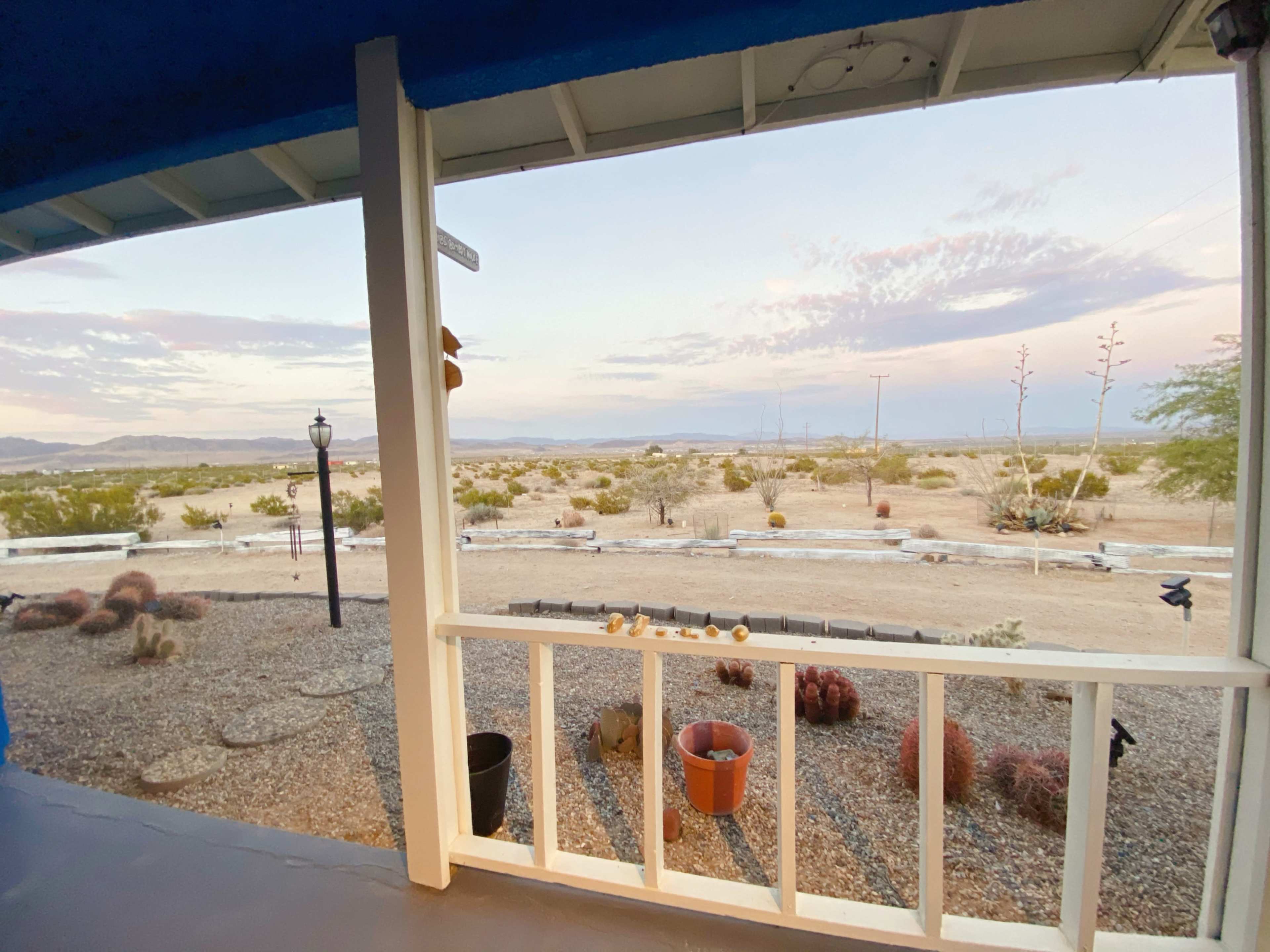 The image shows a view from a porch overlooking a desert landscape with sparse vegetation and distant mountains under a colorful sky.