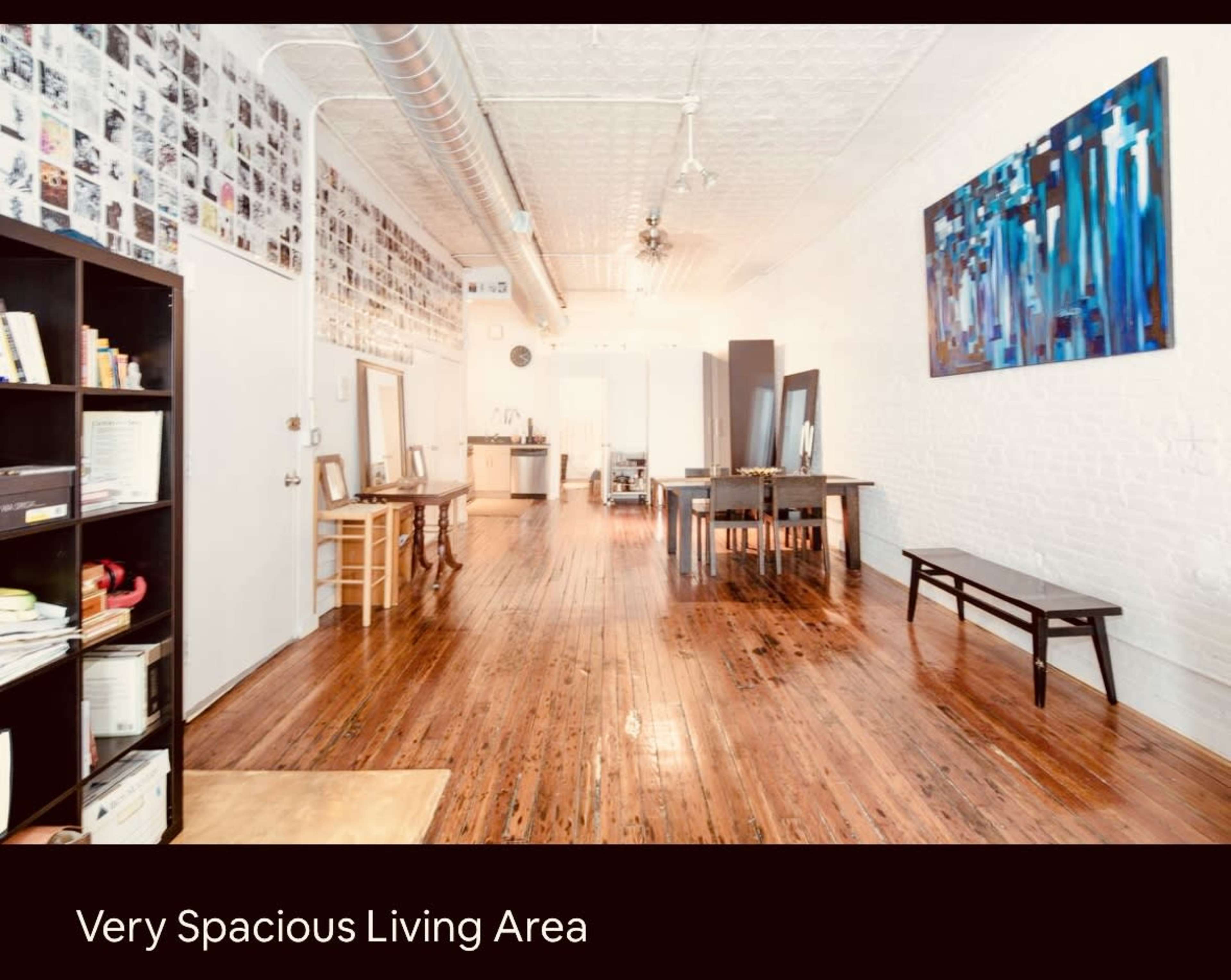 The image shows a large living area with polished wooden floors, white brick walls, and a wall adorned with numerous black-and-white photographs.