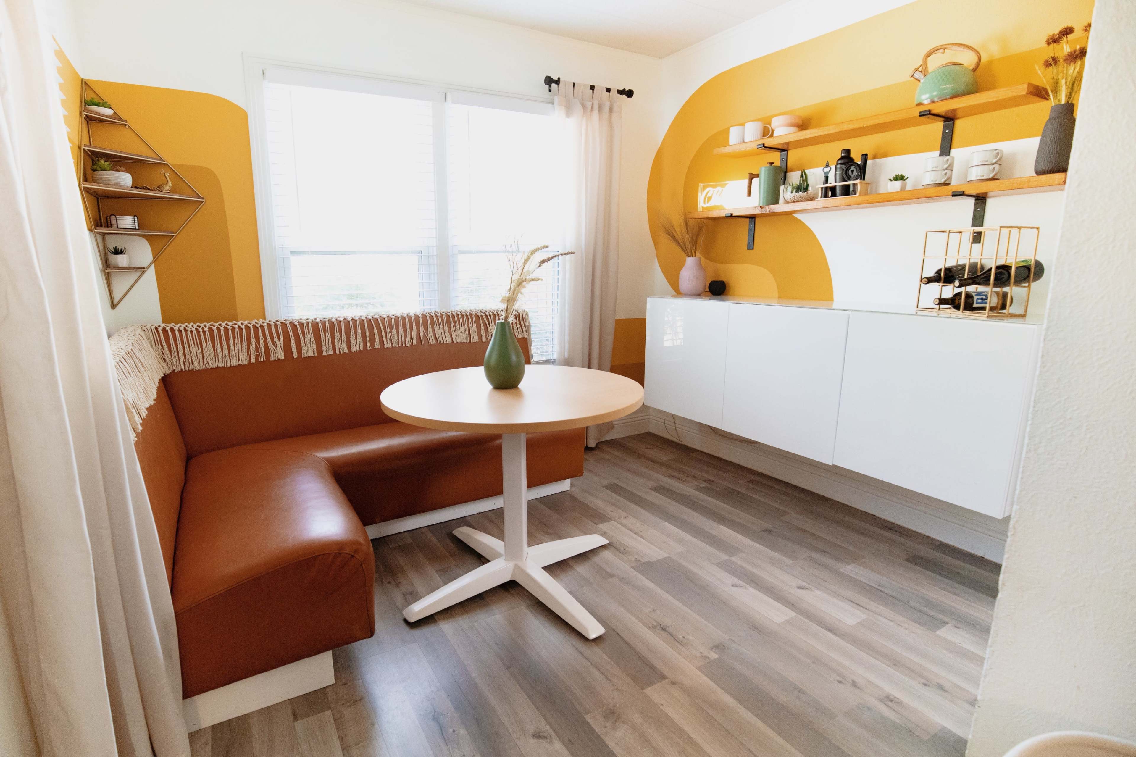 A cozy dining nook features a round table next to a brown upholstered bench and white cabinetry with decorative shelving.