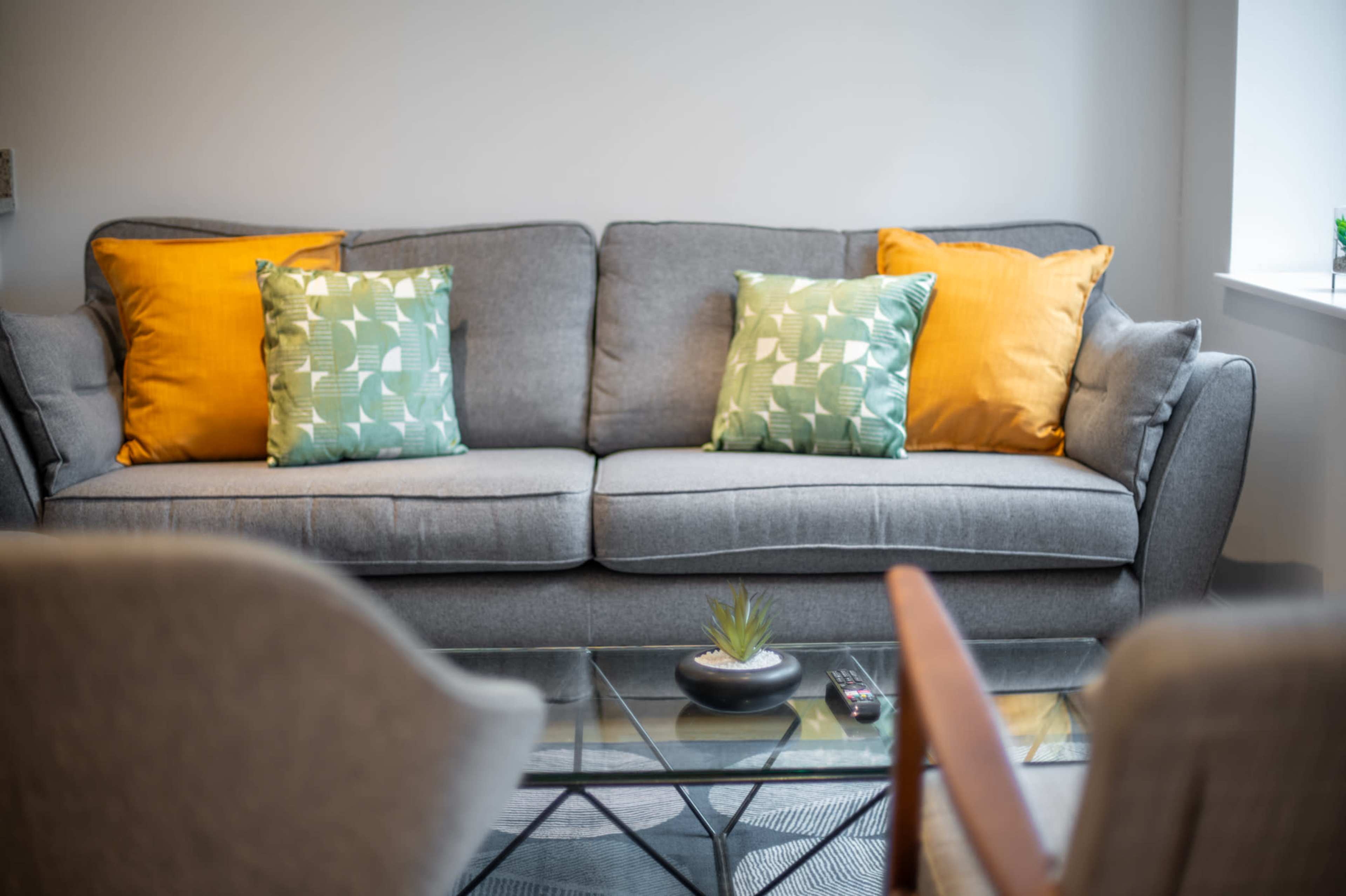 A gray sofa with green and yellow decorative pillows is positioned in front of a glass coffee table, which holds a small plant.