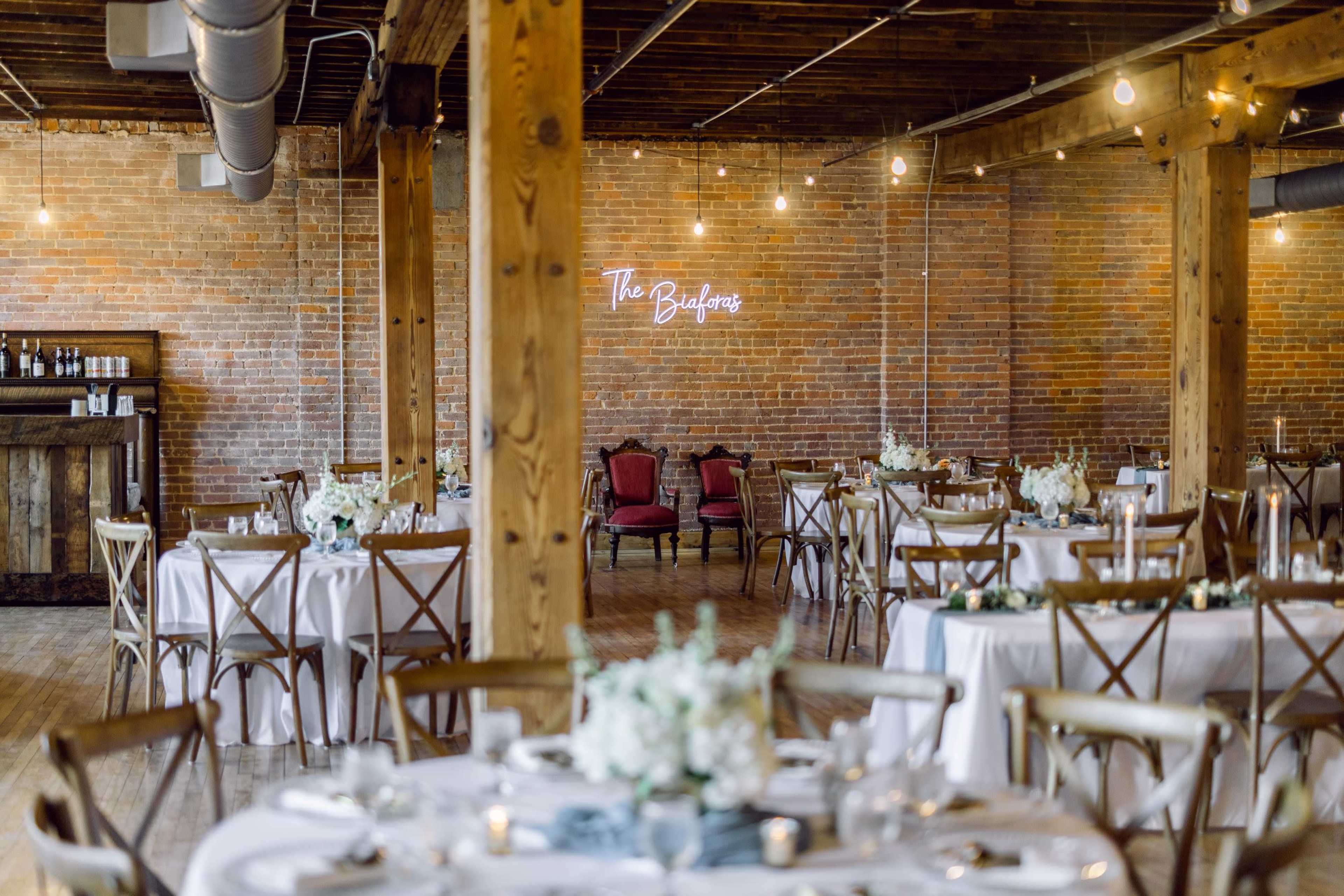 A spacious event venue with wooden beams, round tables set with white tablecloths, and a neon sign that reads "The Building."
