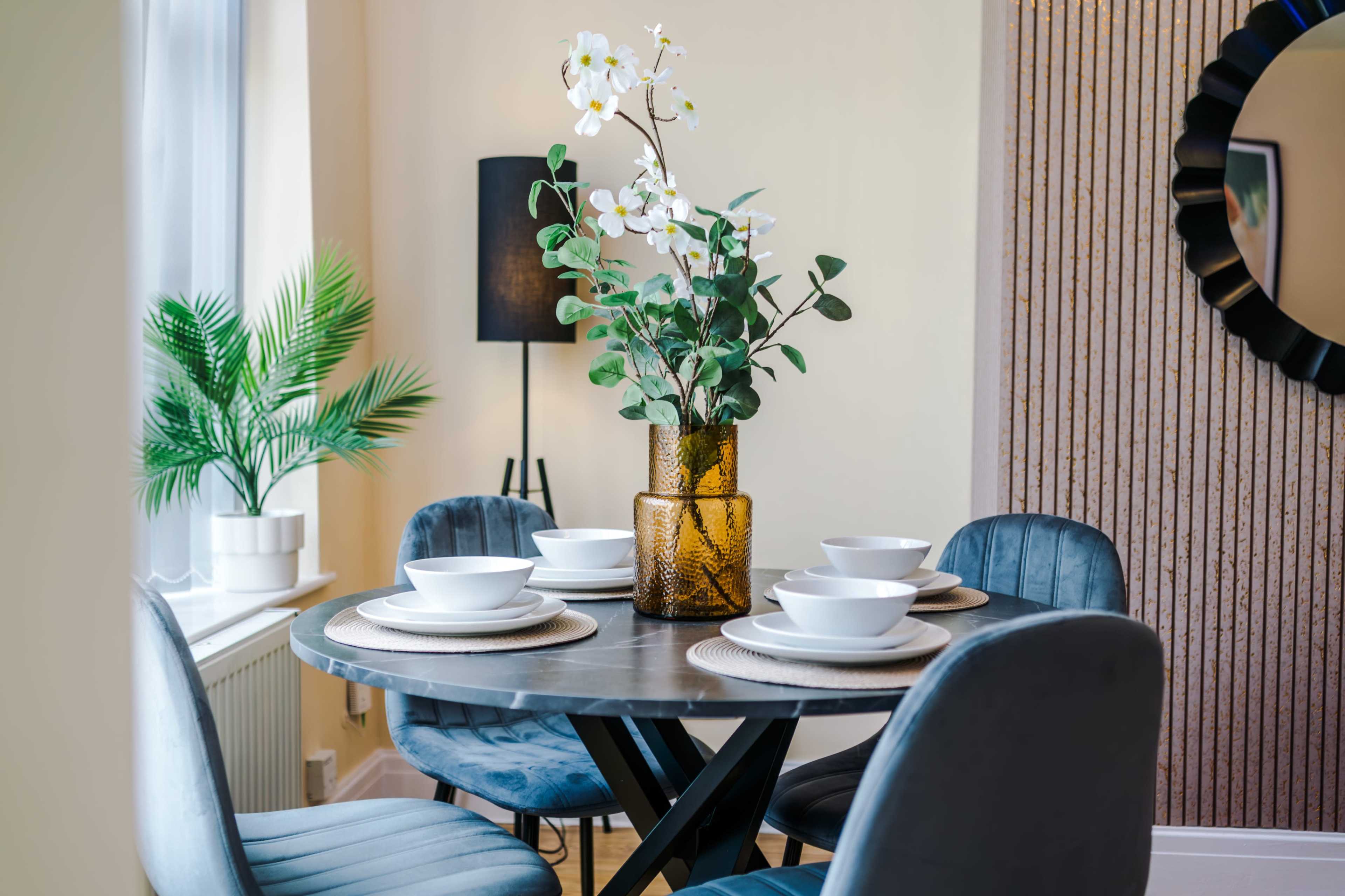 A round dining table set with white plates and a vase of flowers is surrounded by upholstered chairs in a bright room featuring a plant and a mirror.