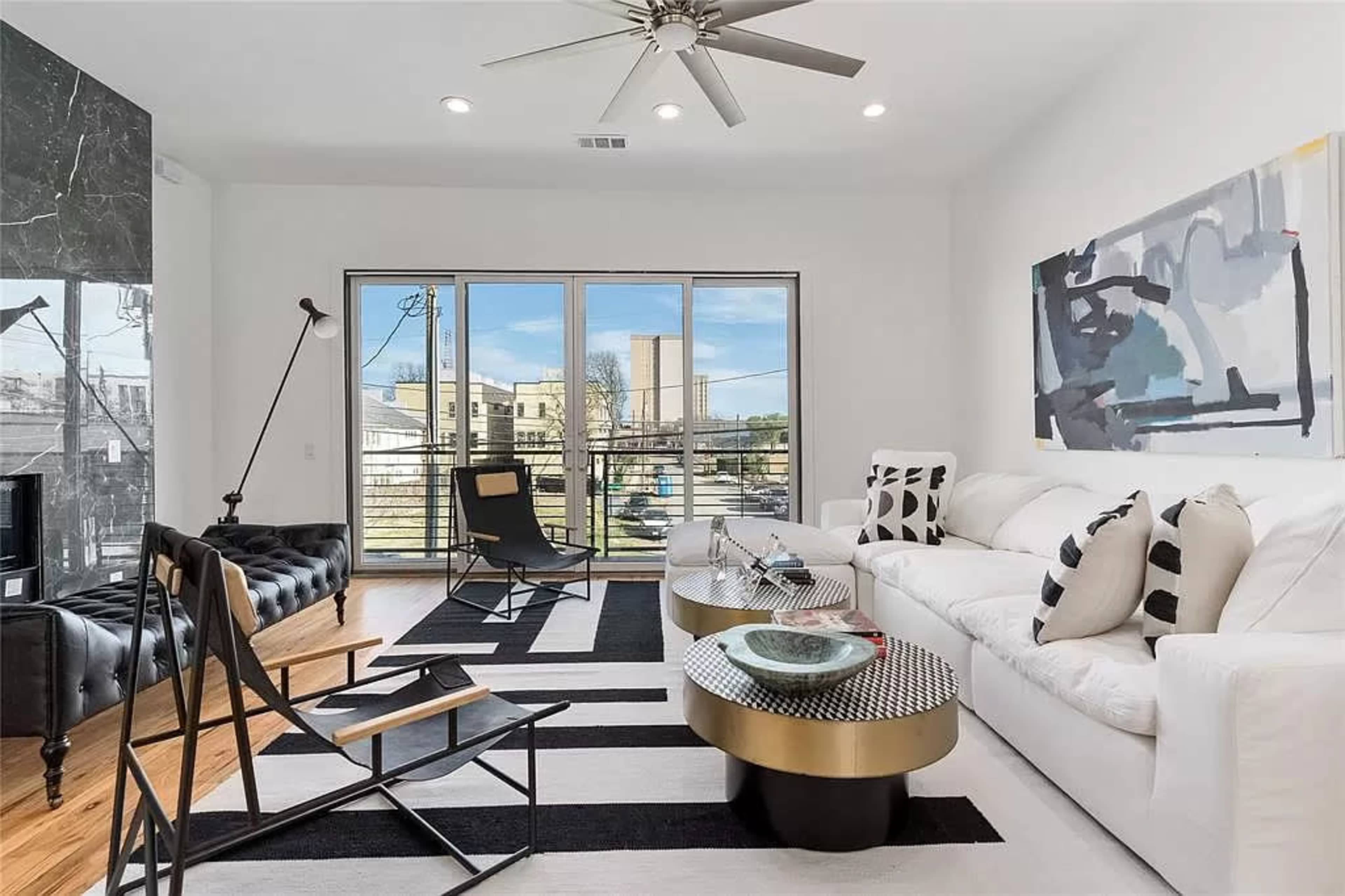 A modern living room featuring a white sofa, black and white patterned rug, and large windows that provide outdoor views.