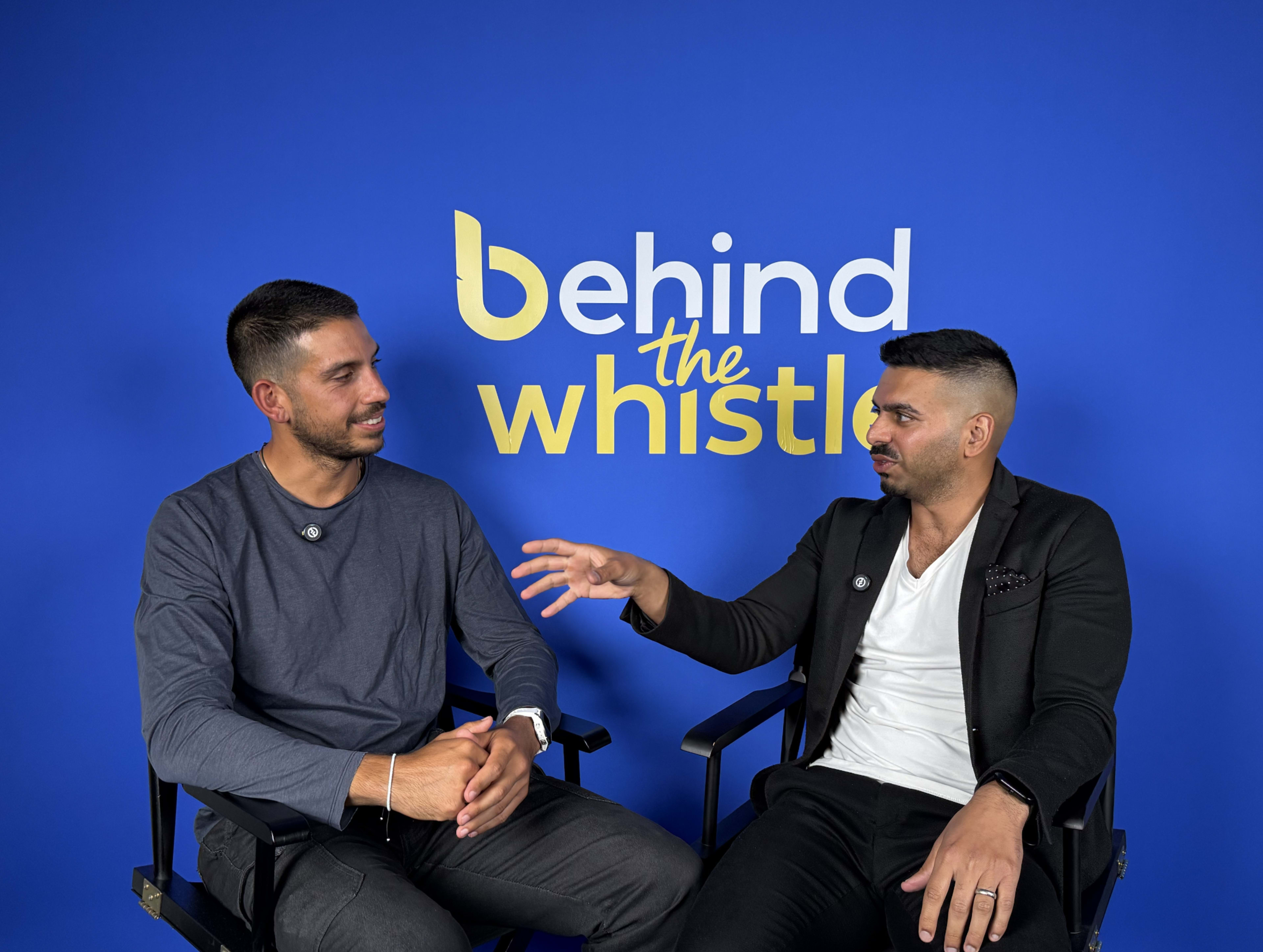 Two men are seated in director's chairs in front of a blue backdrop with the text "behind the whistle."