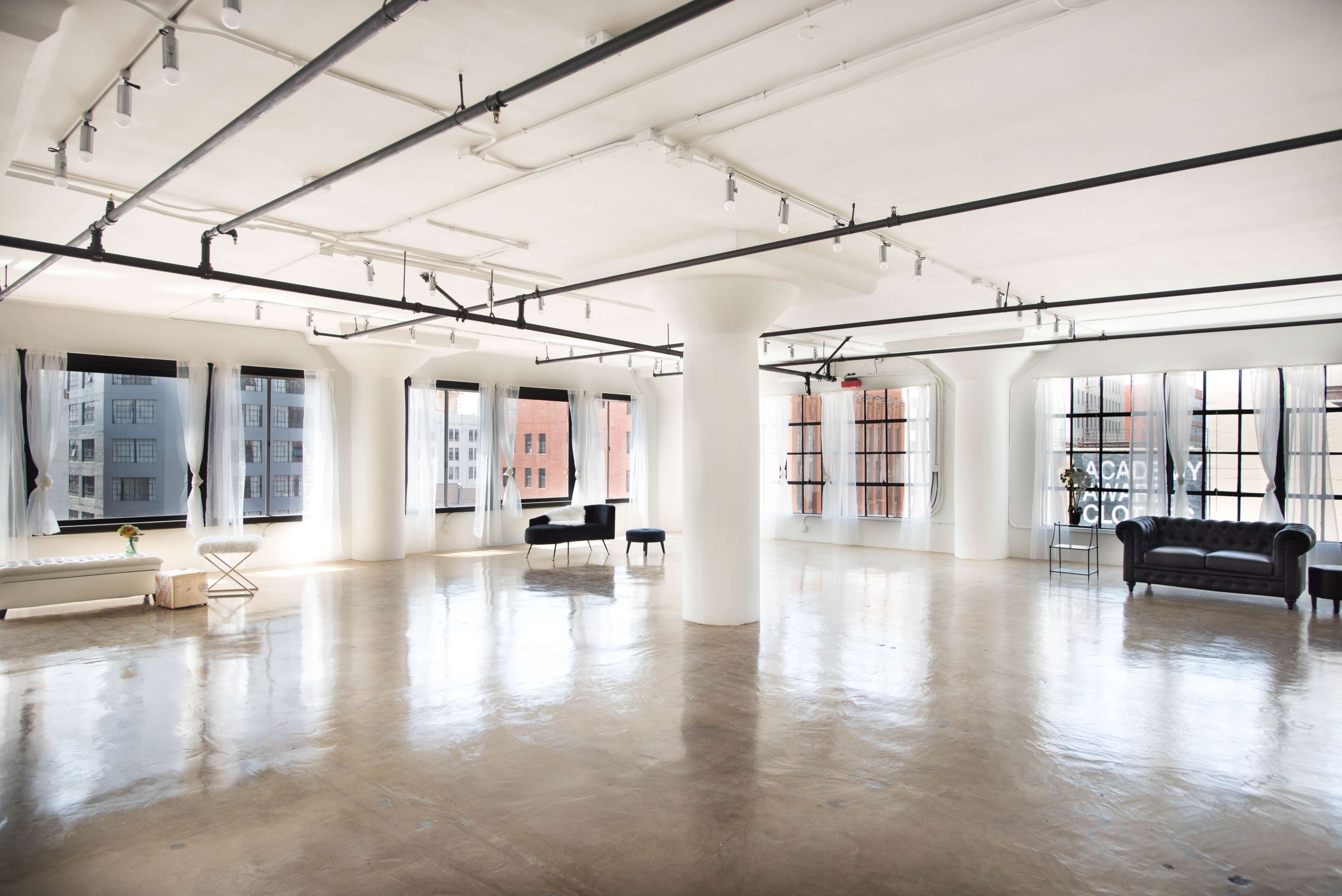 A spacious, empty room features large windows, polished concrete floors, and minimalist furniture arranged around its central support pillars.