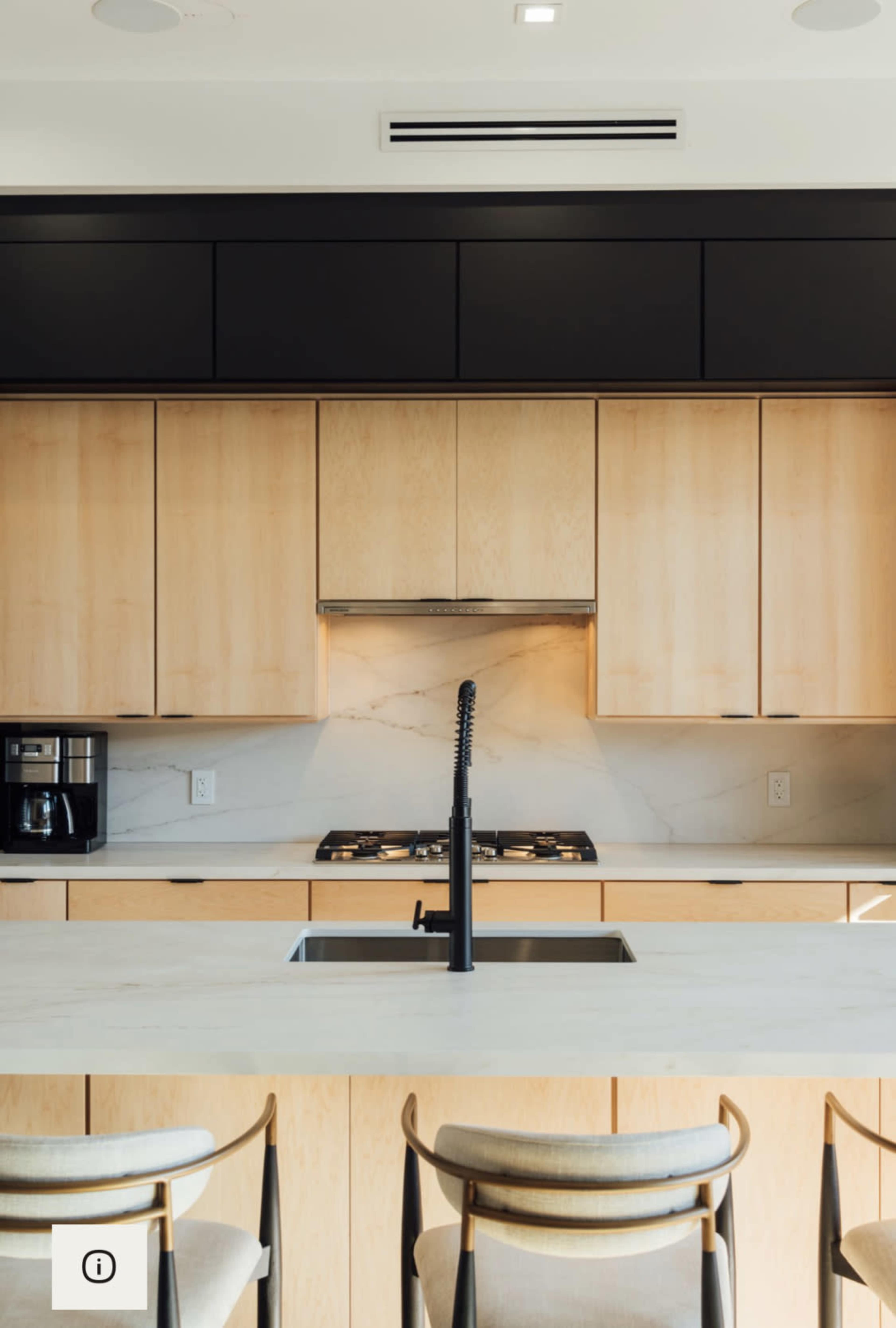 A modern kitchen with wooden cabinets, a marble backsplash, a black sink and faucet, and two stools at a light-colored countertop.