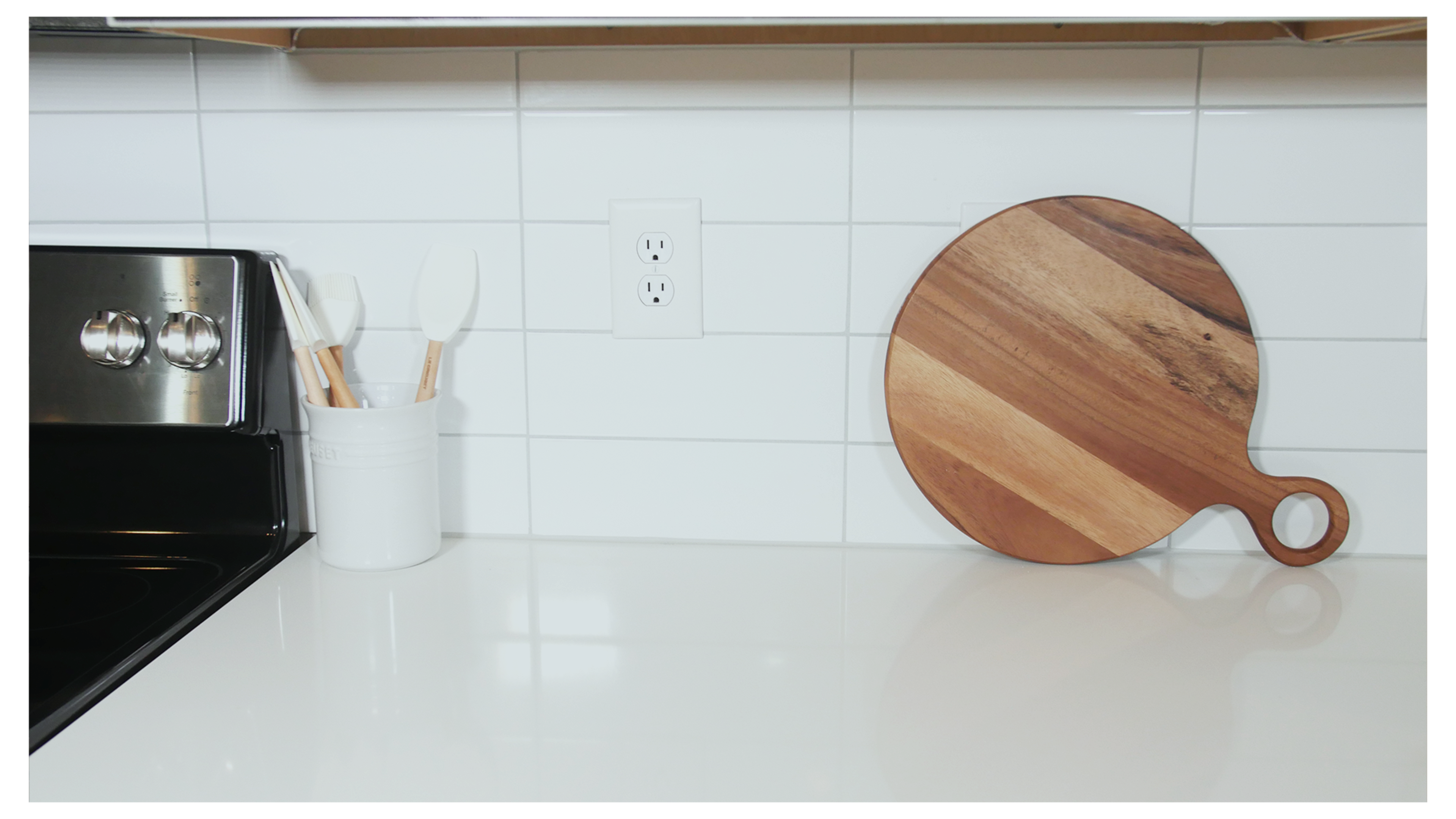 The image shows a kitchen countertop with a black stove, a white container holding utensils, and a wooden cutting board resting against the wall.