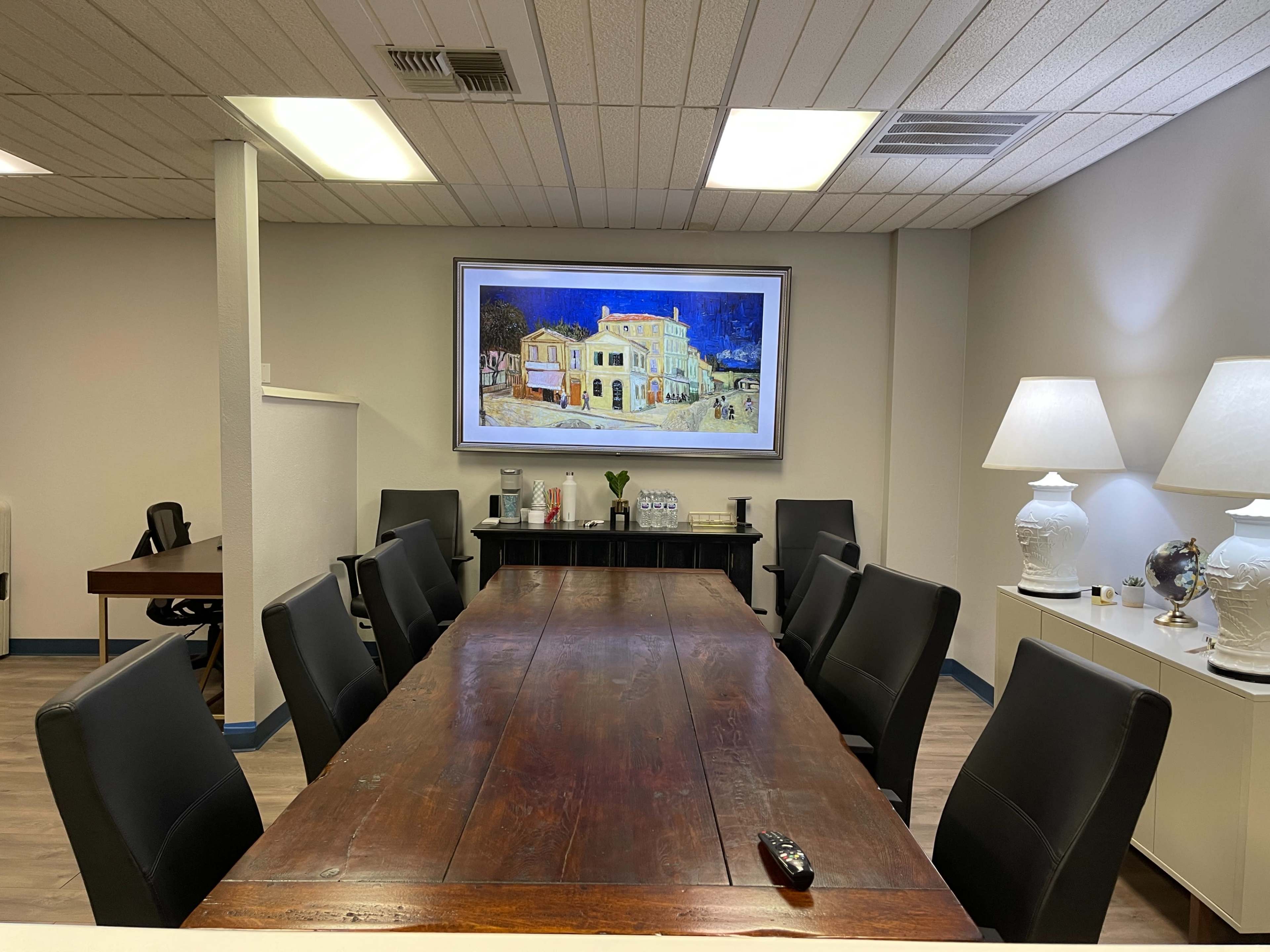 A long wooden conference table is set in a meeting room with black chairs, framed by a large painting and two lamps on opposite sides.