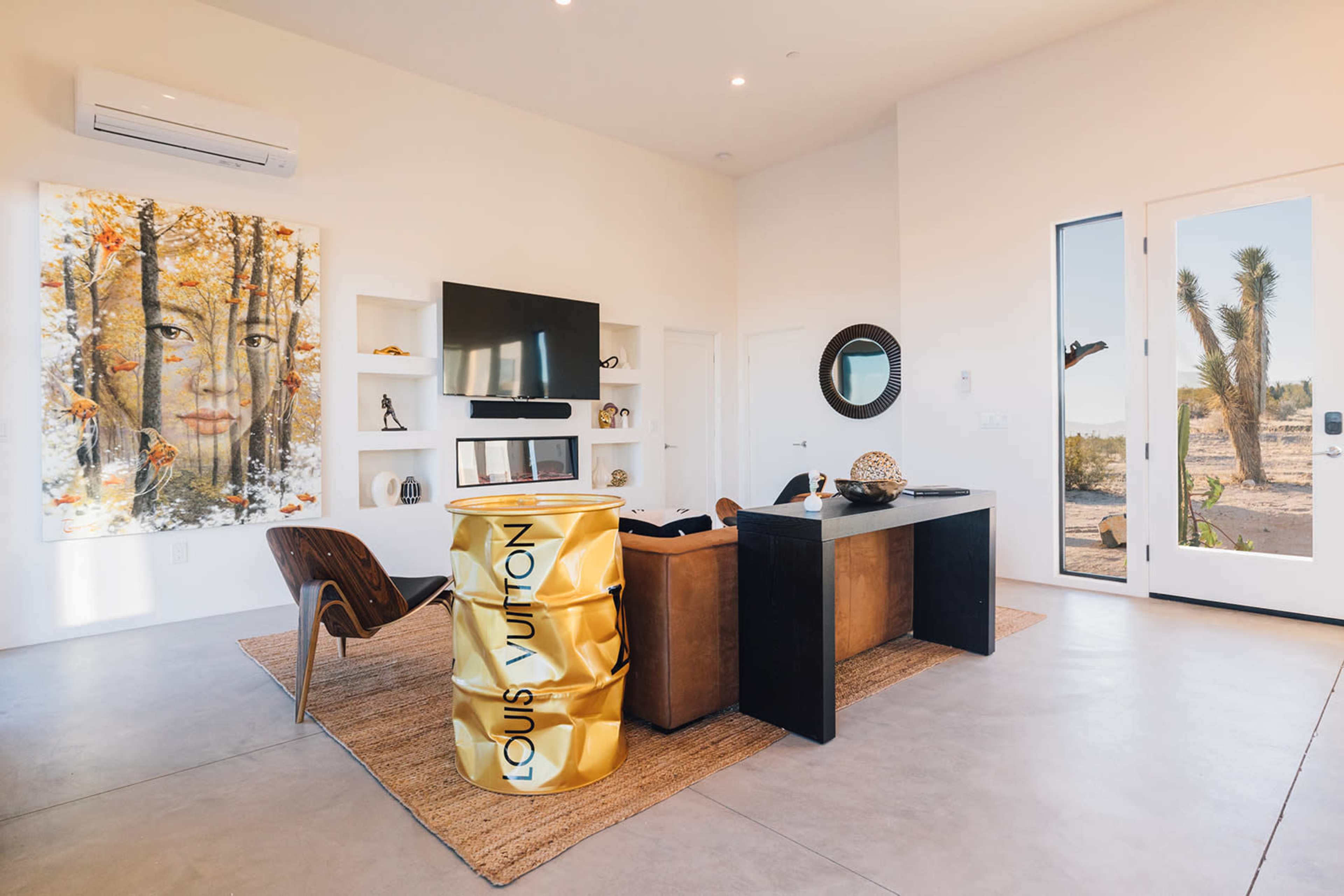 The scene features a modern living room with a large artwork of a face, a television on a wall-mounted shelf, a stylish office desk, and a large gold Louis Vuitton accessory, with a view of desert landscaping through a glass door.