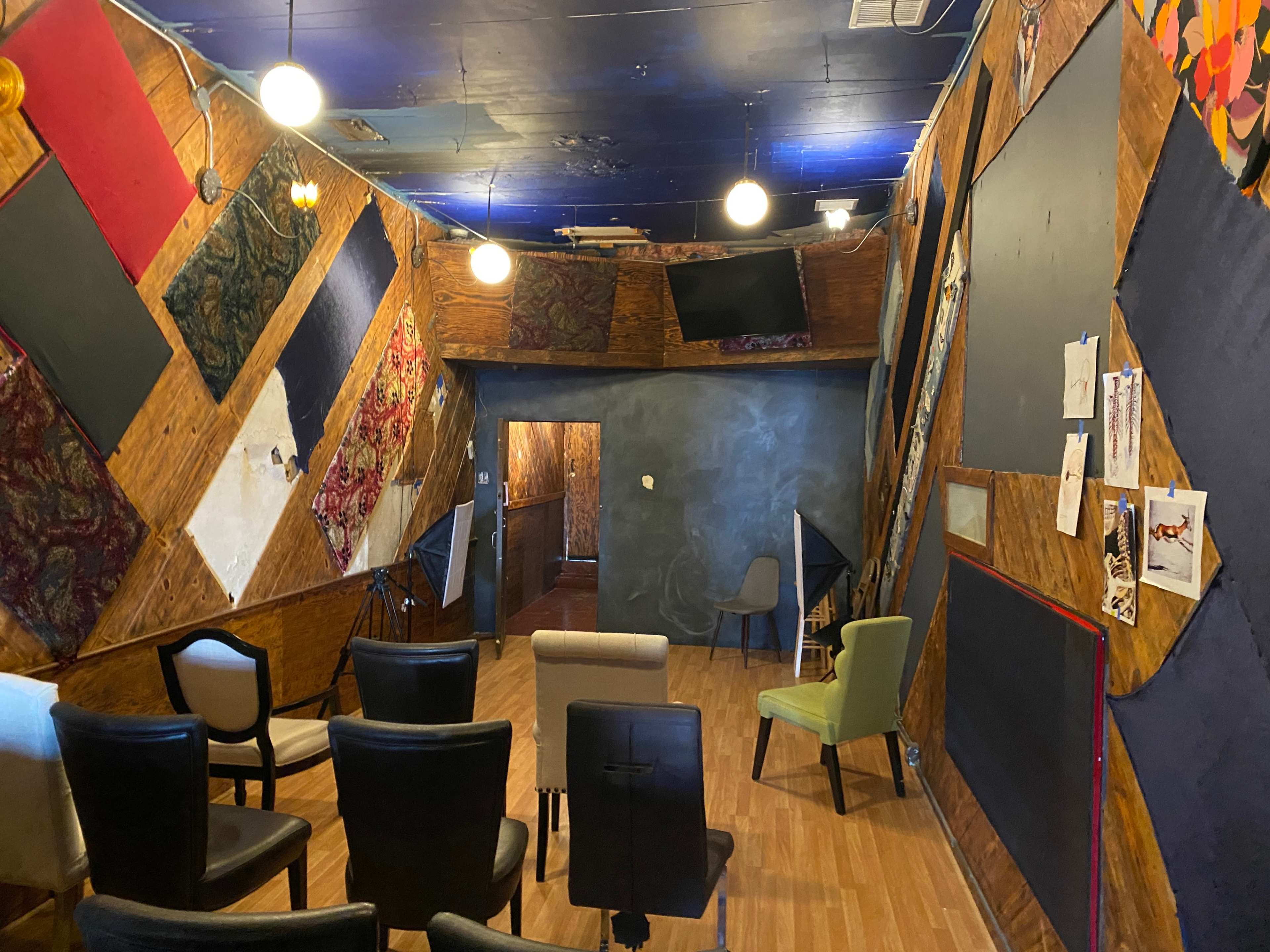 A room with a unique, textured wall design featuring various colors and patterns, arranged seating in rows, and a dark area intended for presentations or performances.
