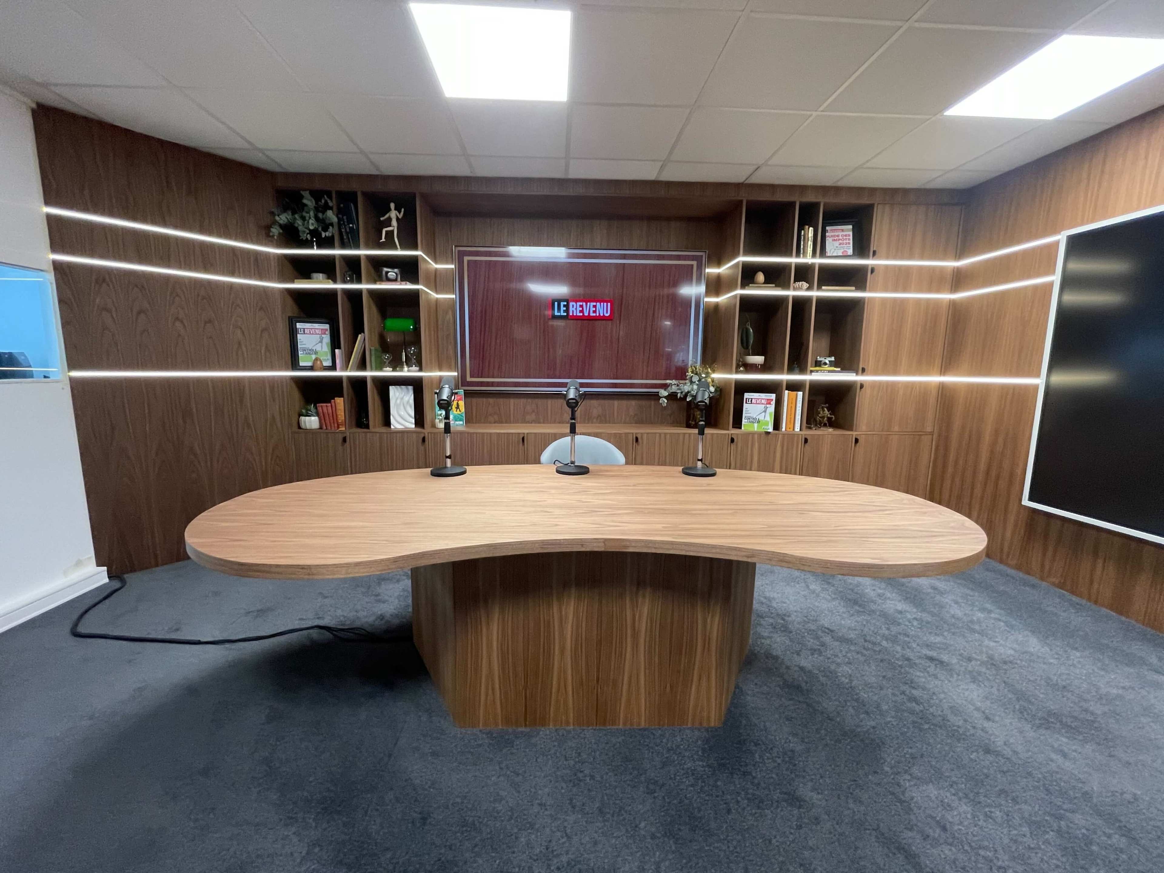 The image shows a modern studio setup with a curved wooden desk, two microphones, and a backdrop featuring shelving with decorative items and a screen displaying "LE BREV."