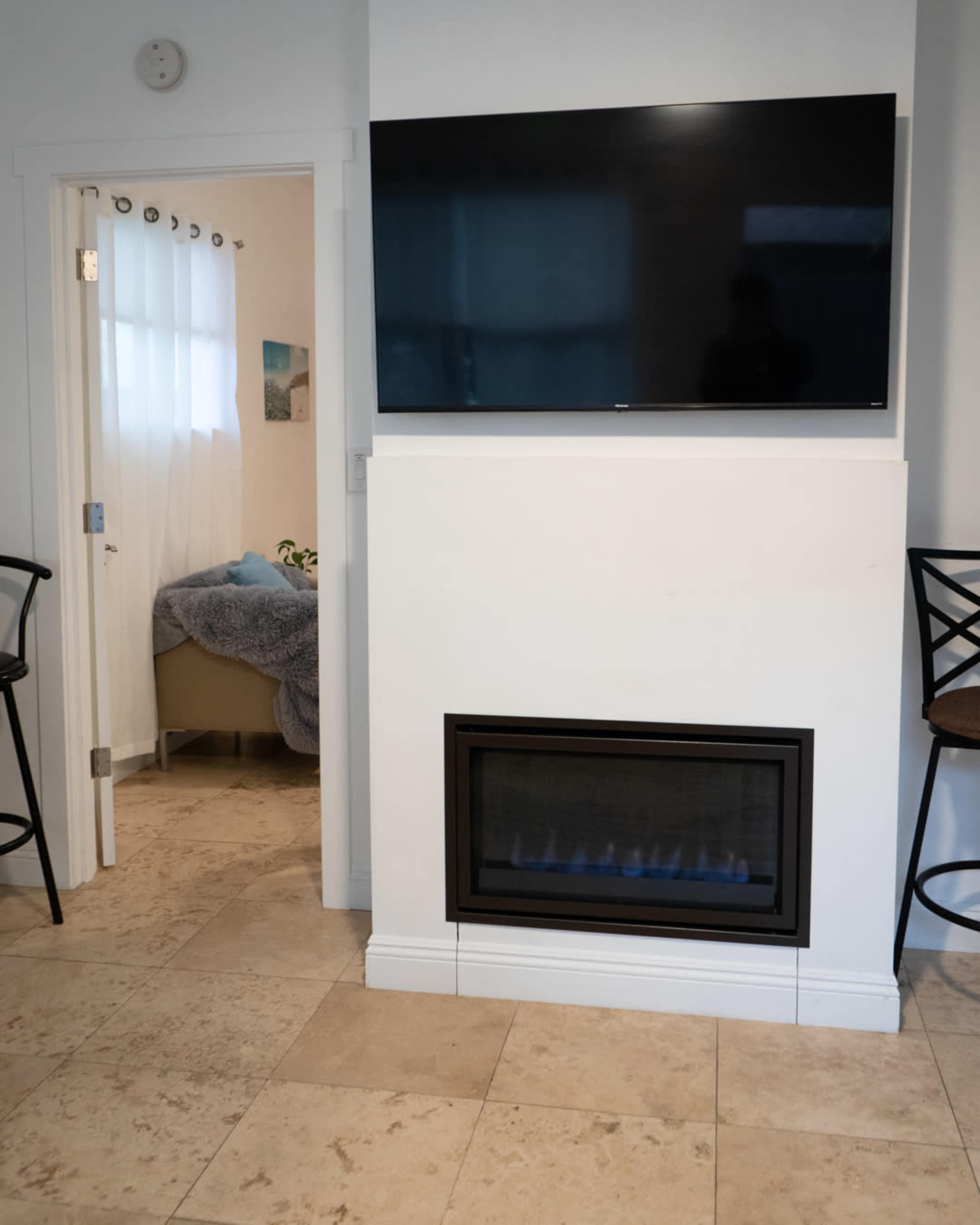 The image shows a modern living space with a wall-mounted television above a built-in fireplace, and a view of a doorway leading to a room with a bed.
