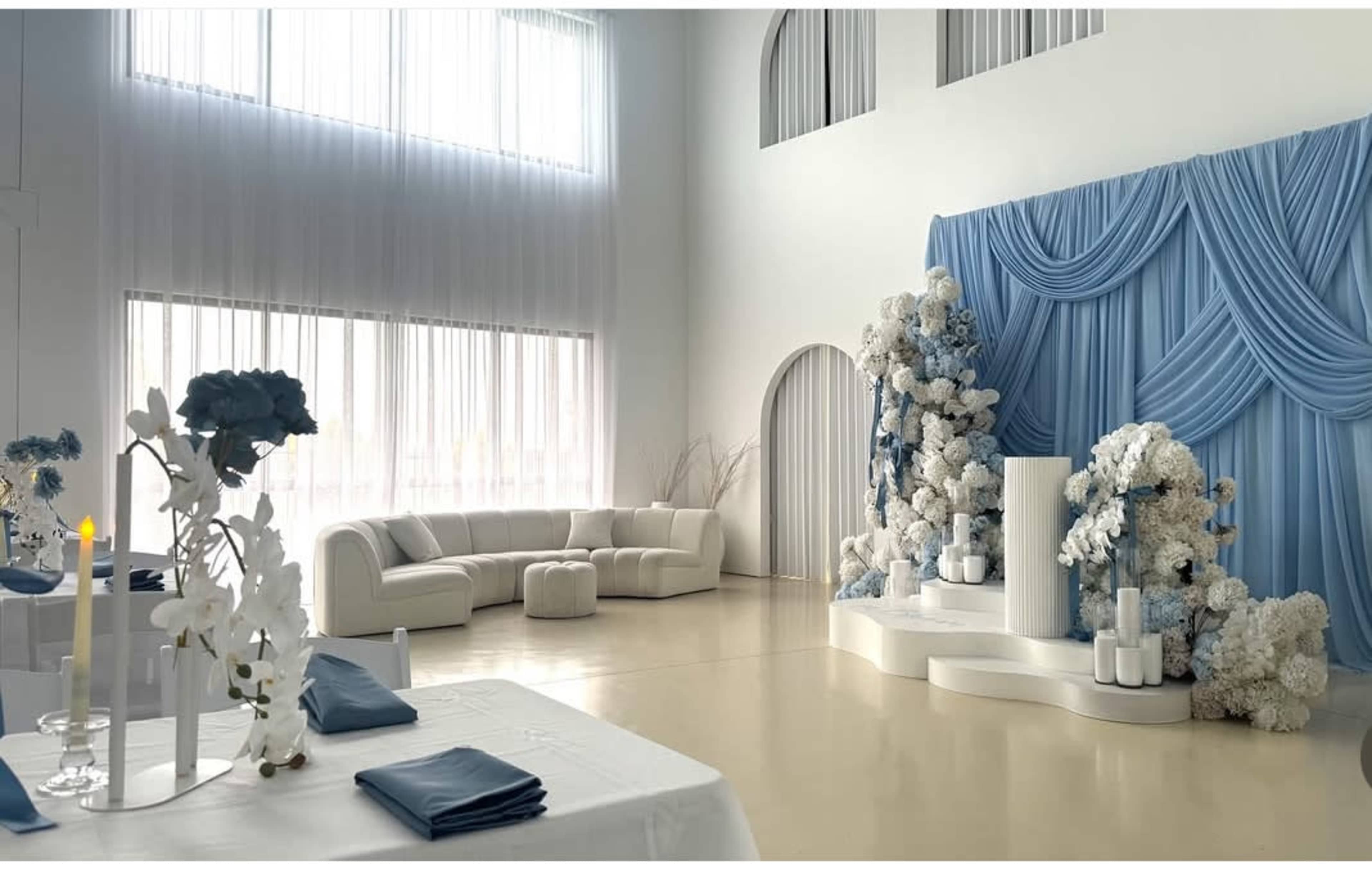 The image shows a modern event space featuring a light-colored sofa, tables set with blue napkins, and a decorative backdrop with blue drapes and floral arrangements.