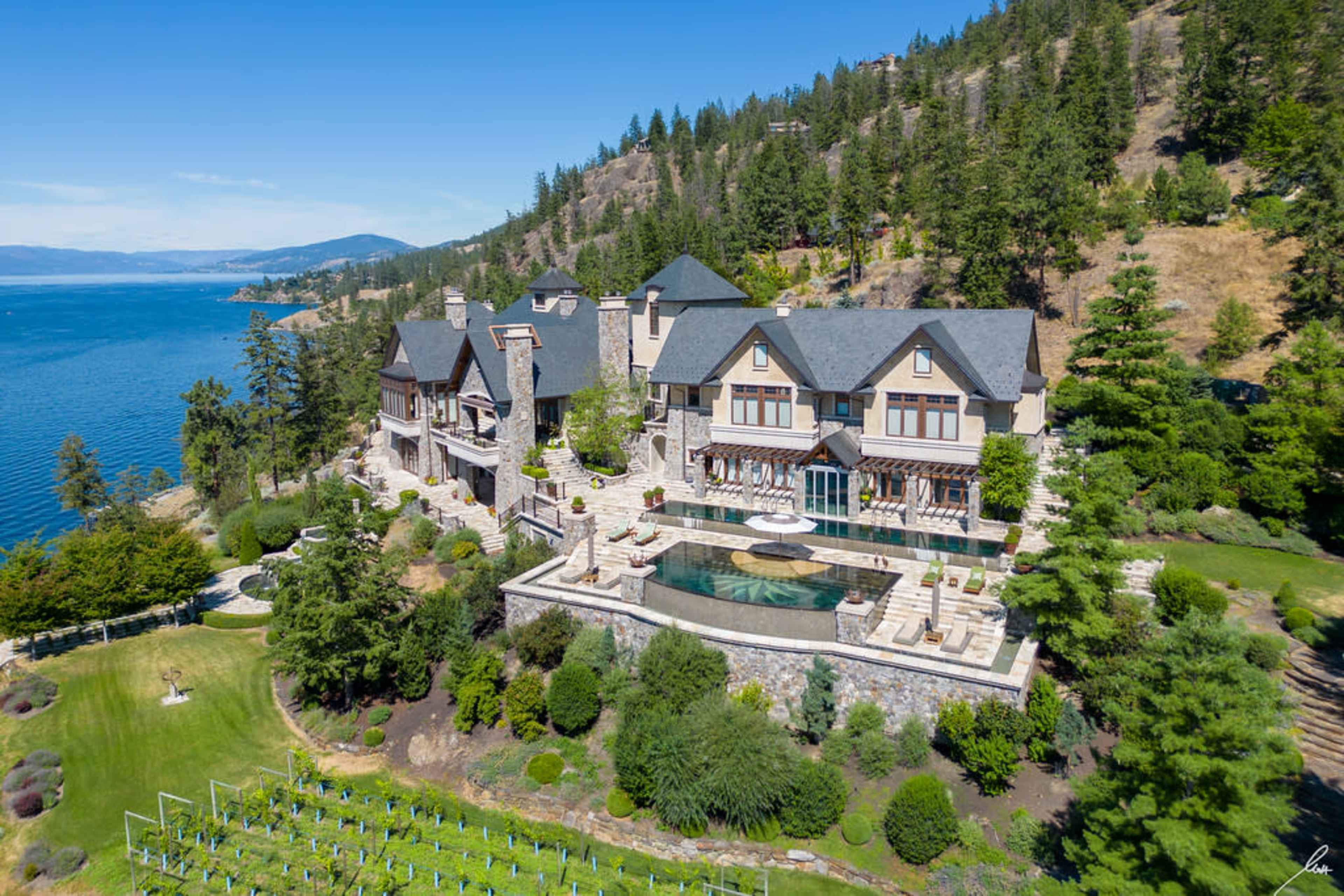 A large, stone-finished mansion with multiple gables overlooks a serene lake, surrounded by lush greenery and landscaped gardens.