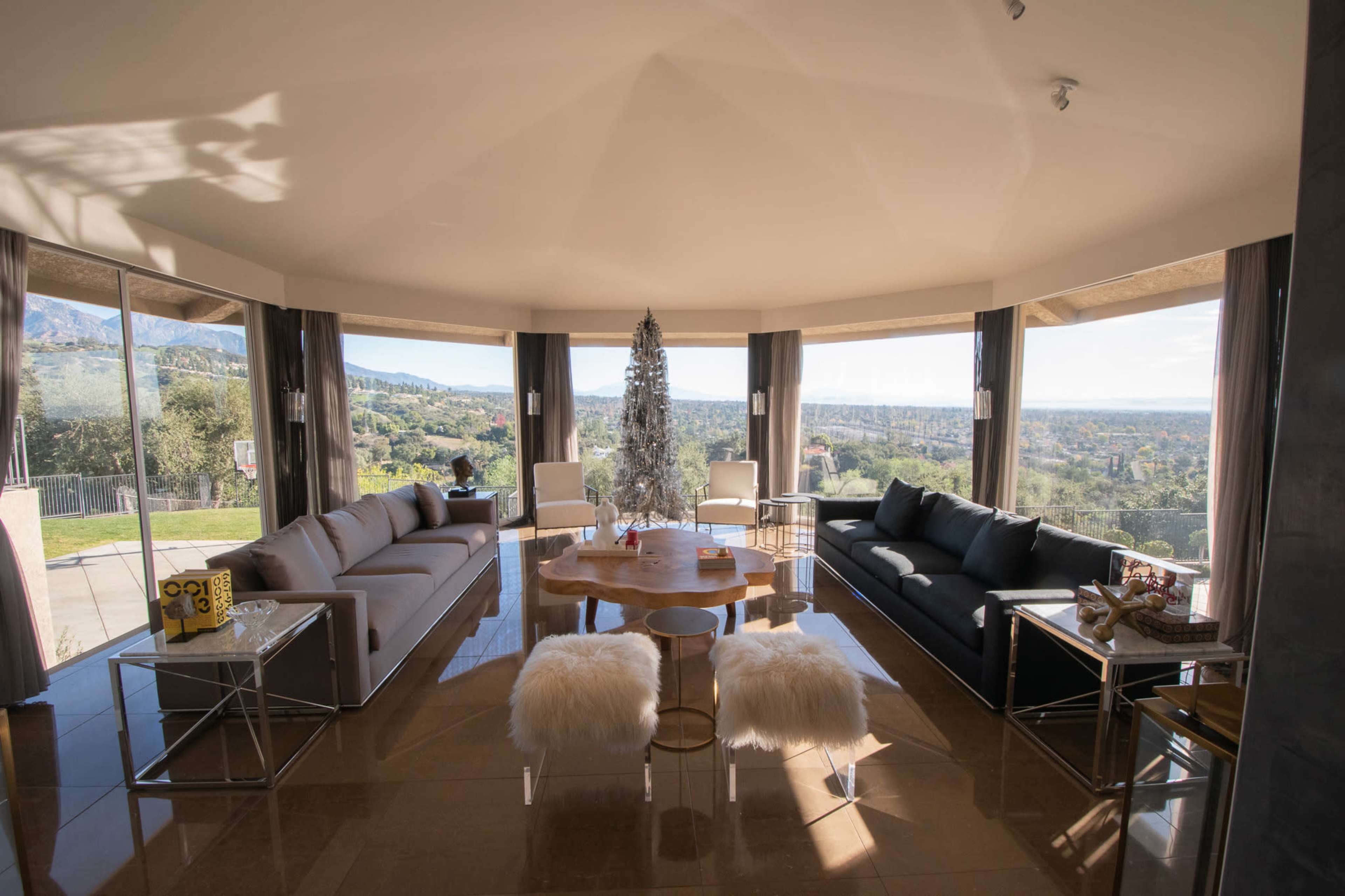 The image shows a modern living room with large windows offering a scenic view, featuring a round table surrounded by various seating arrangements and a decorative tree.