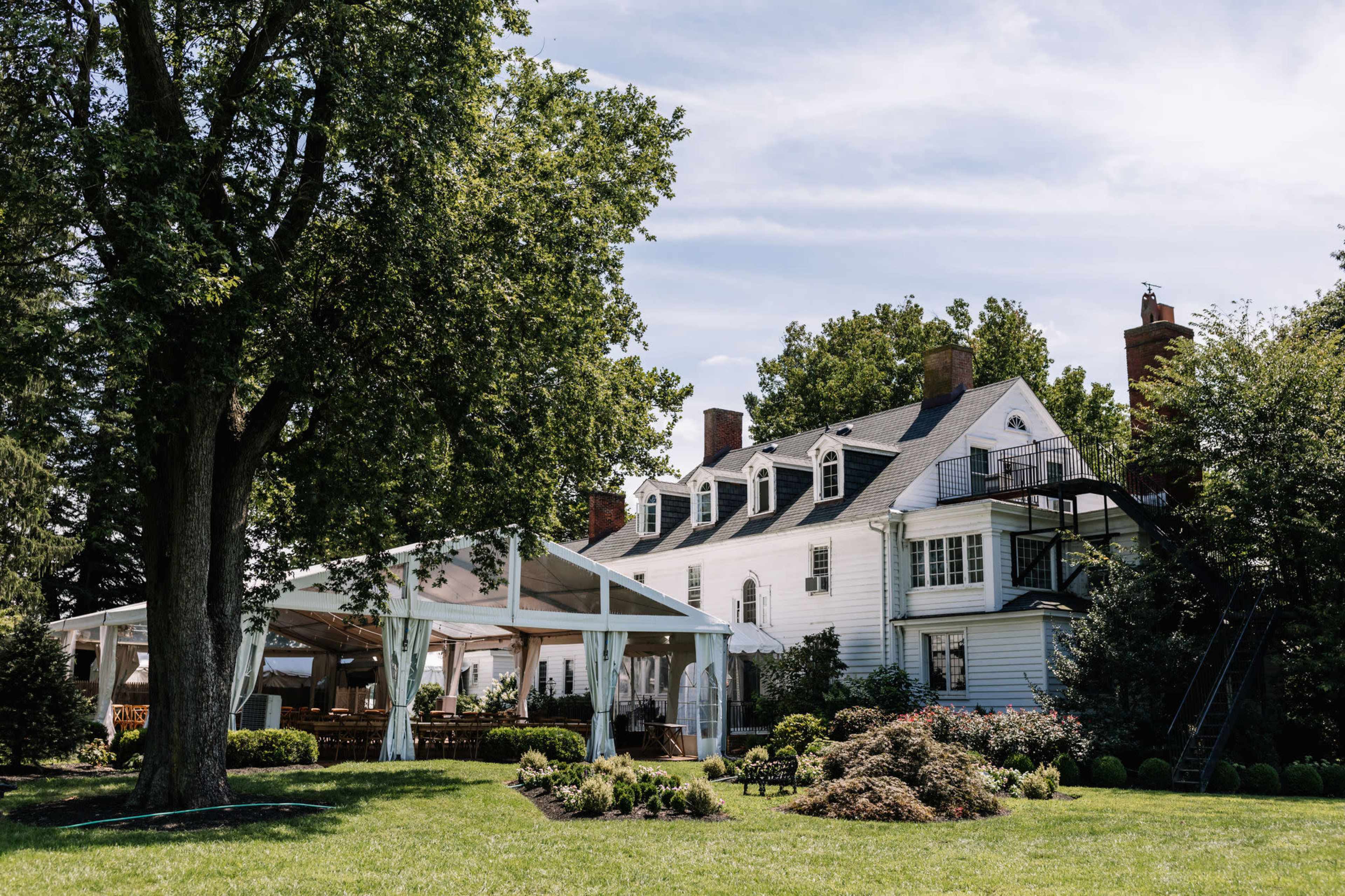A large white house with multiple gabled roofs and a spacious tented area in the foreground, set against a backdrop of green trees and shrubs.