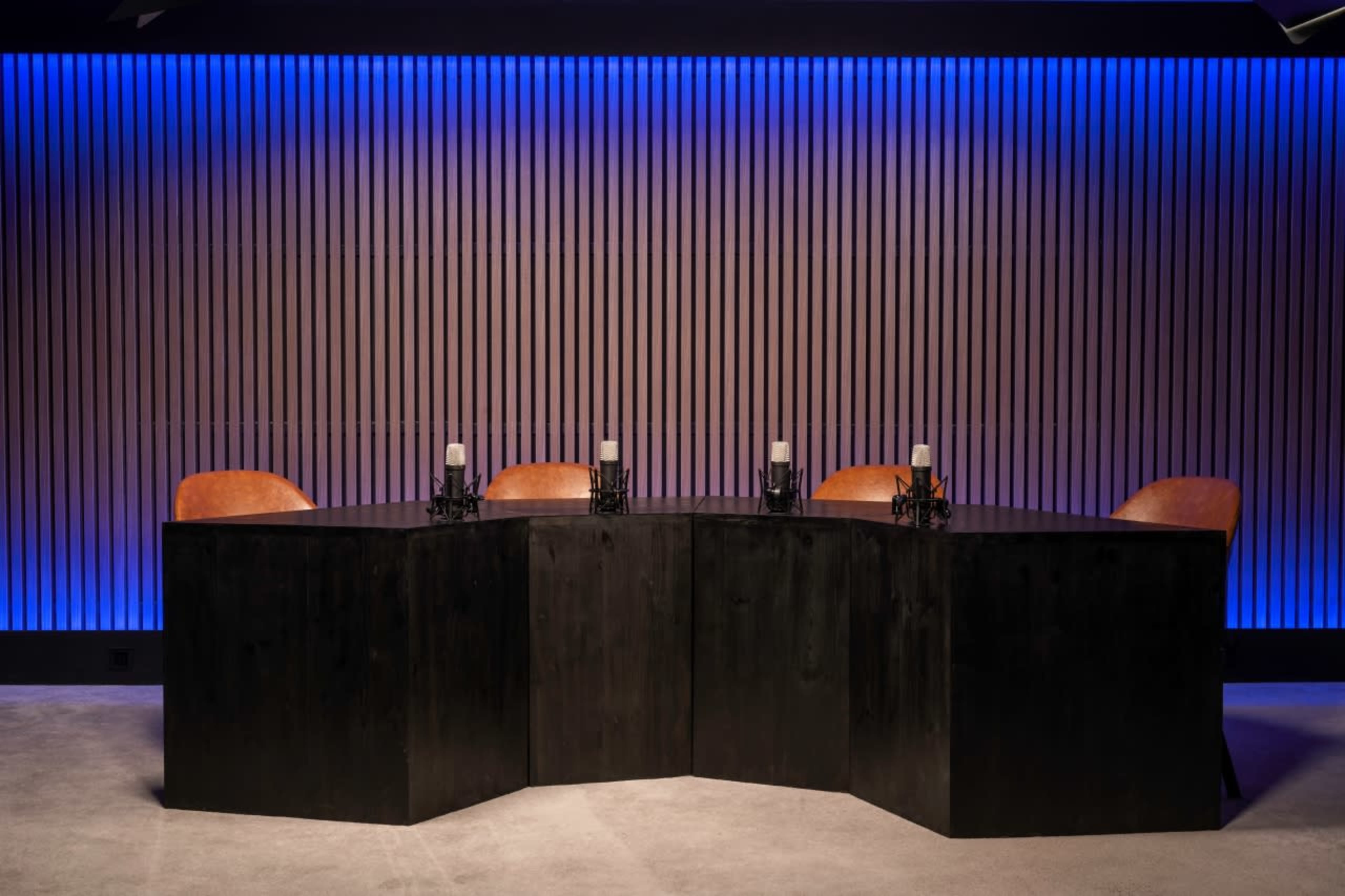 A modern conference table is positioned against a backdrop of vertical wooden slats illuminated with blue lighting, accompanied by four brown leather chairs.