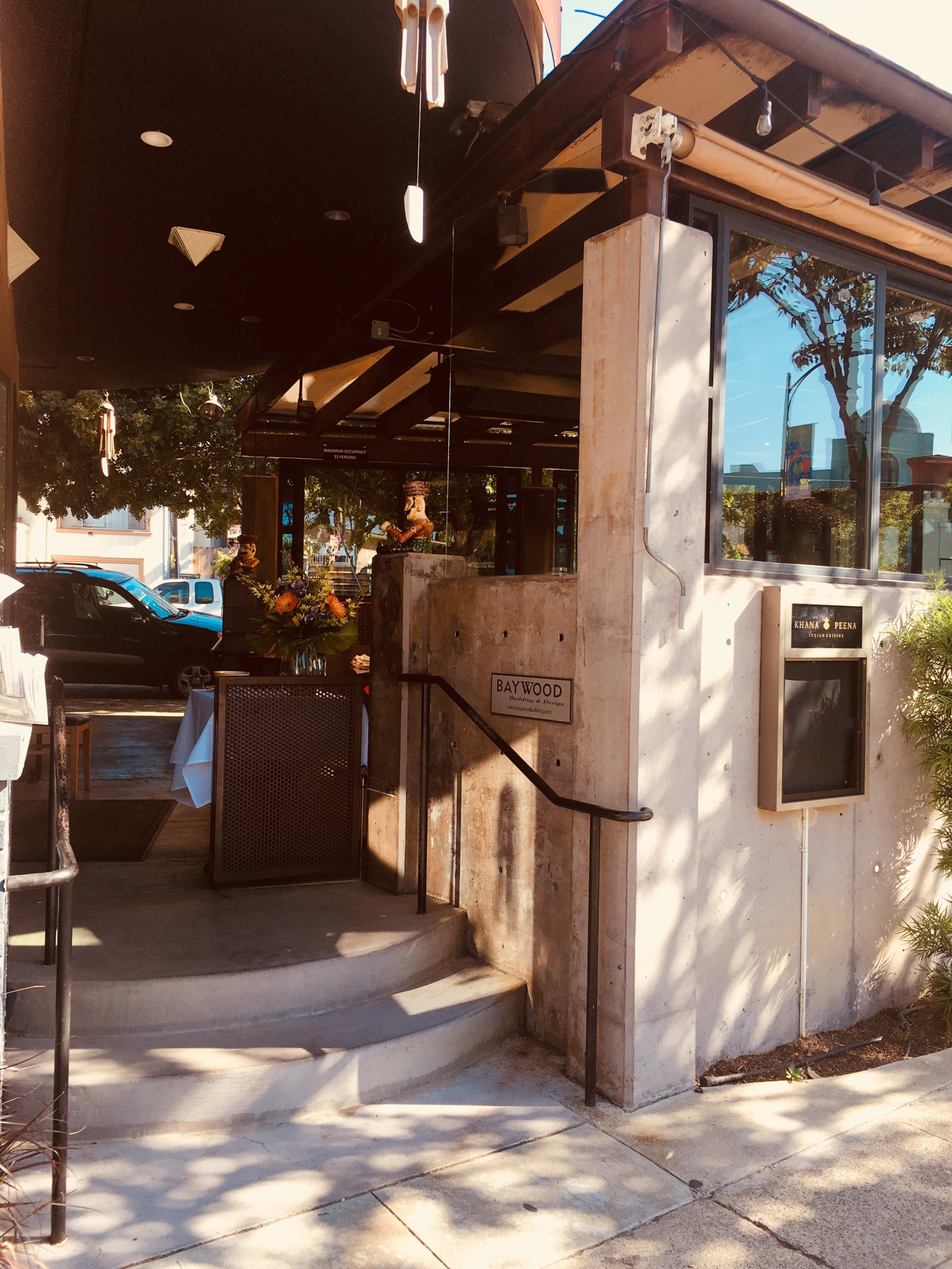 The entrance to a restaurant features a modern concrete and glass design with decorative flowers and a menu display.