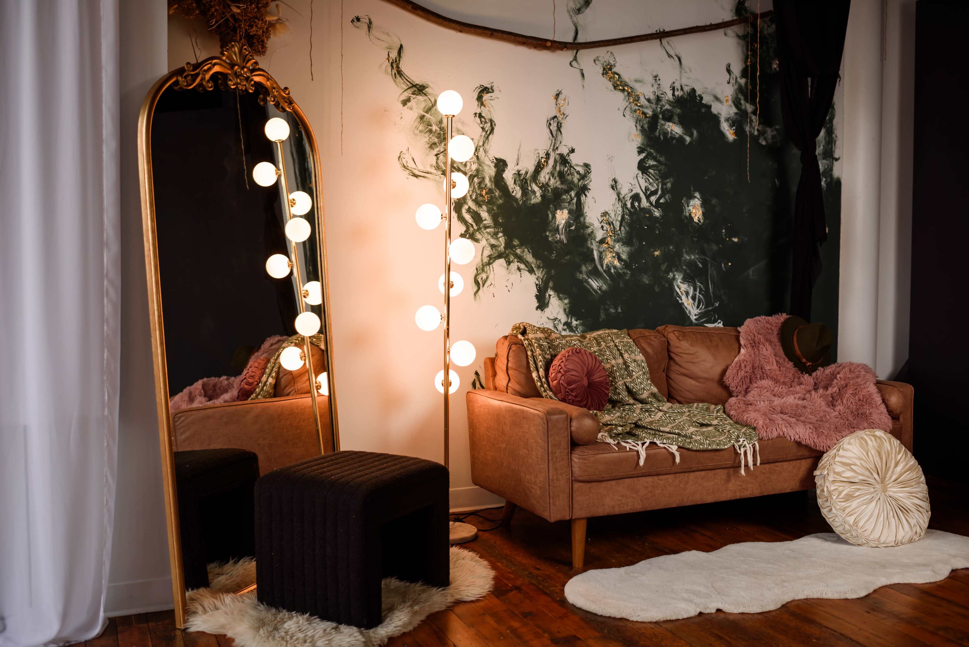 The image features a cozy living room with a brown leather sofa, a tall mirror, and a floor lamp with round bulbs, against a backdrop of an artistically painted wall and wooden flooring.