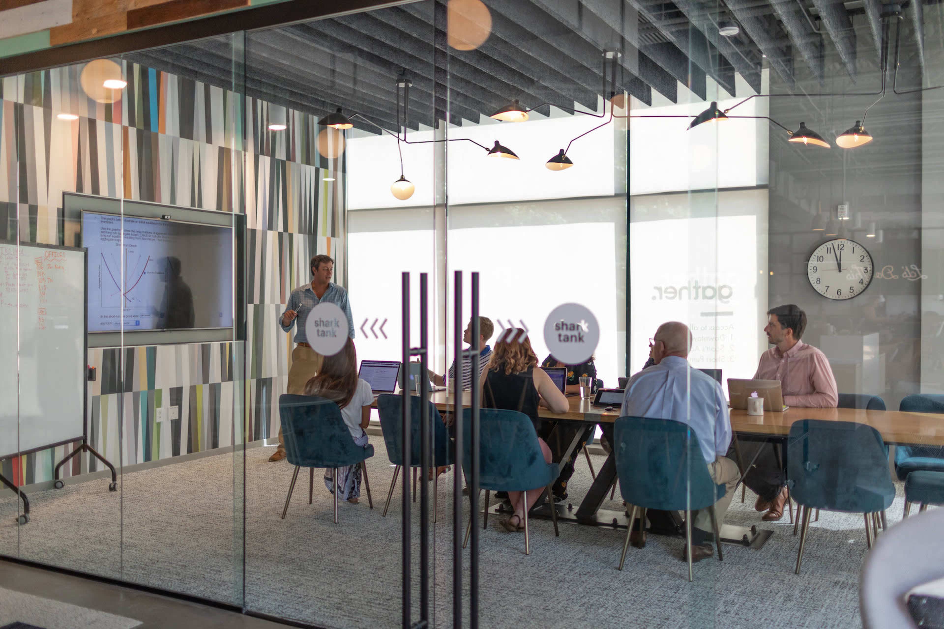 A group of people sit around a conference table in a modern meeting room while a presenter stands by a screen displaying a graph.
