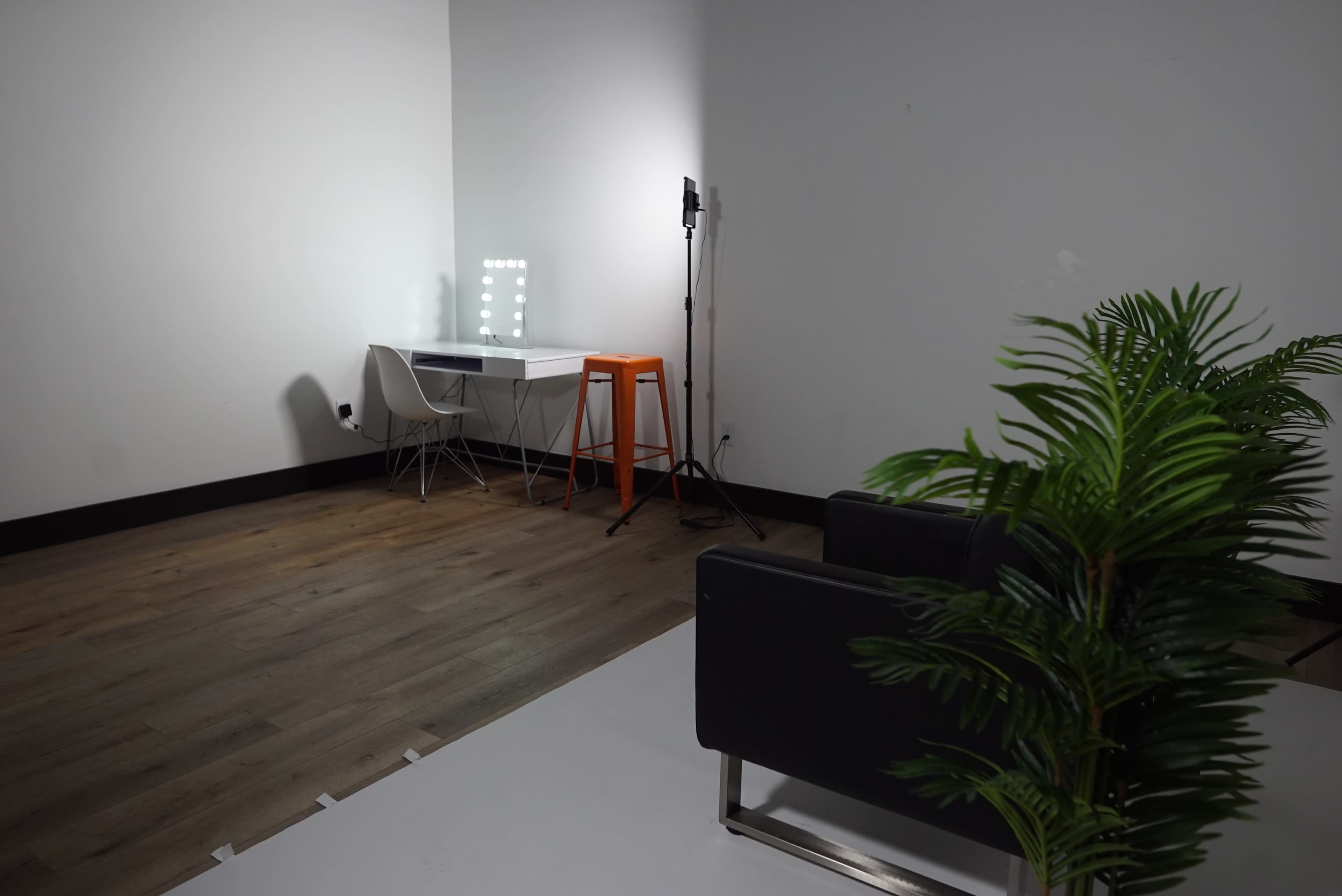 A minimalist room featuring a desk with a lighted mirror, an orange stool, a black sofa, and a potted plant.
