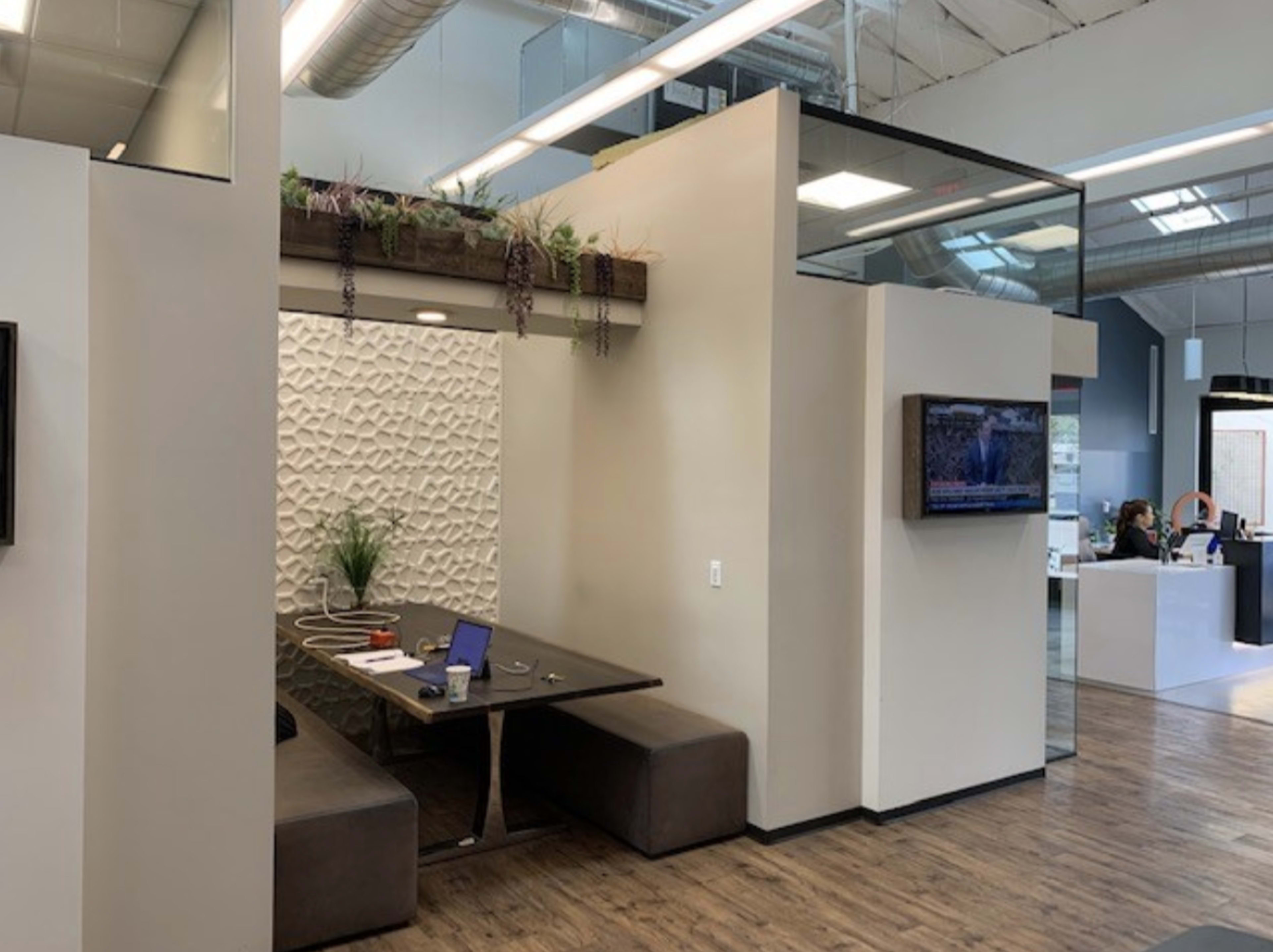 The image shows a modern office space featuring a meeting area with a long table, plush seating, and decorative plants on a partition wall.