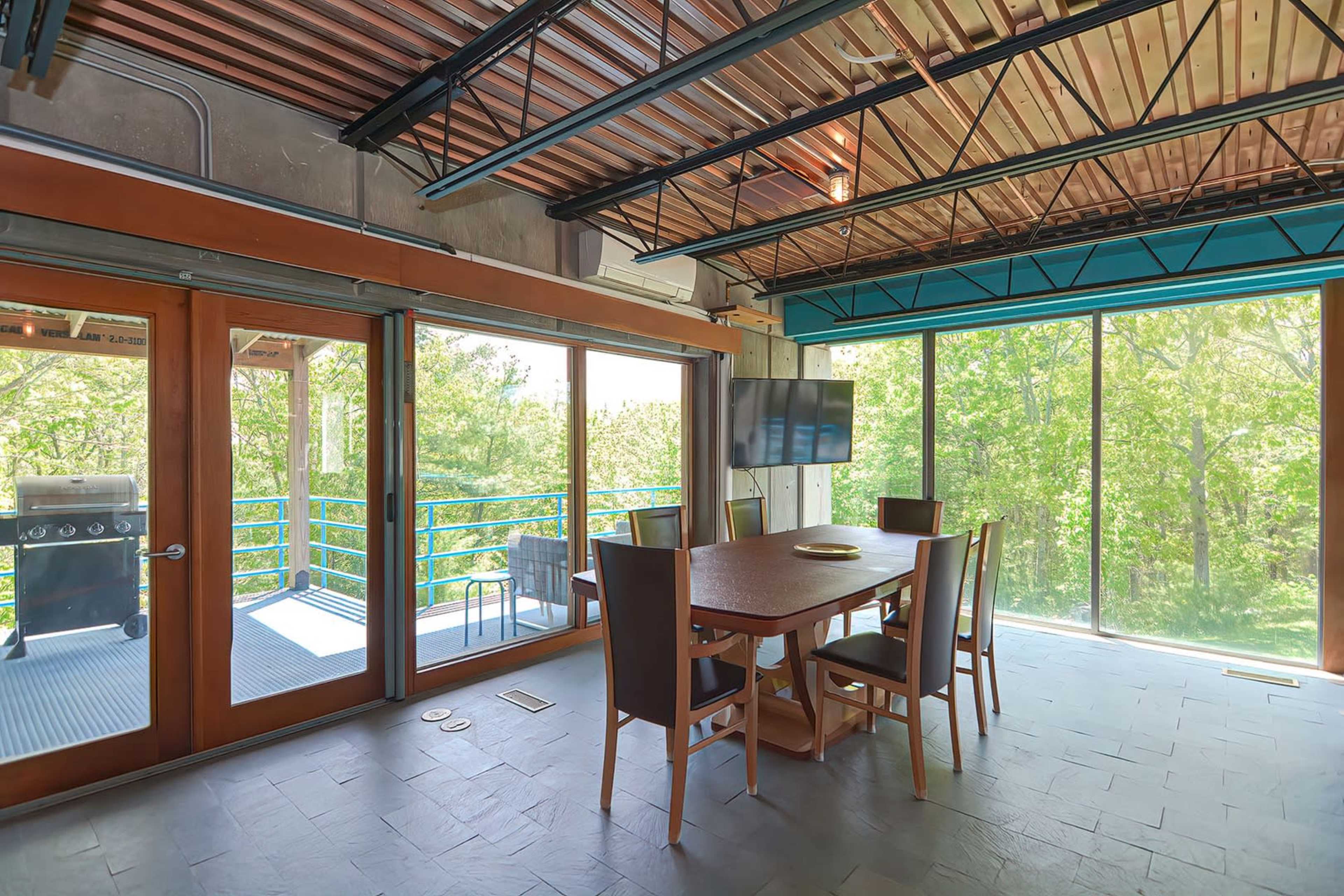 A modern dining area features a wooden table surrounded by chairs, with large windows showcasing a view of trees outside and a patio with a grill.
