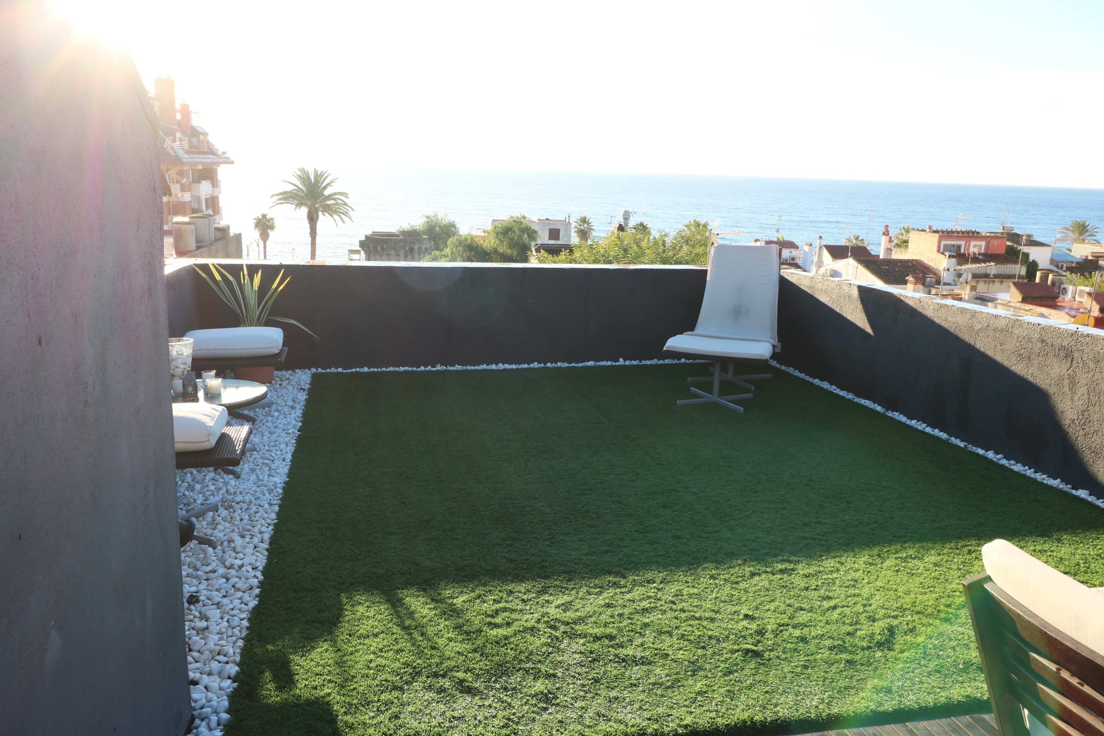 A rooftop terrace features artificial grass, white stones along the edges, and a view of the ocean.