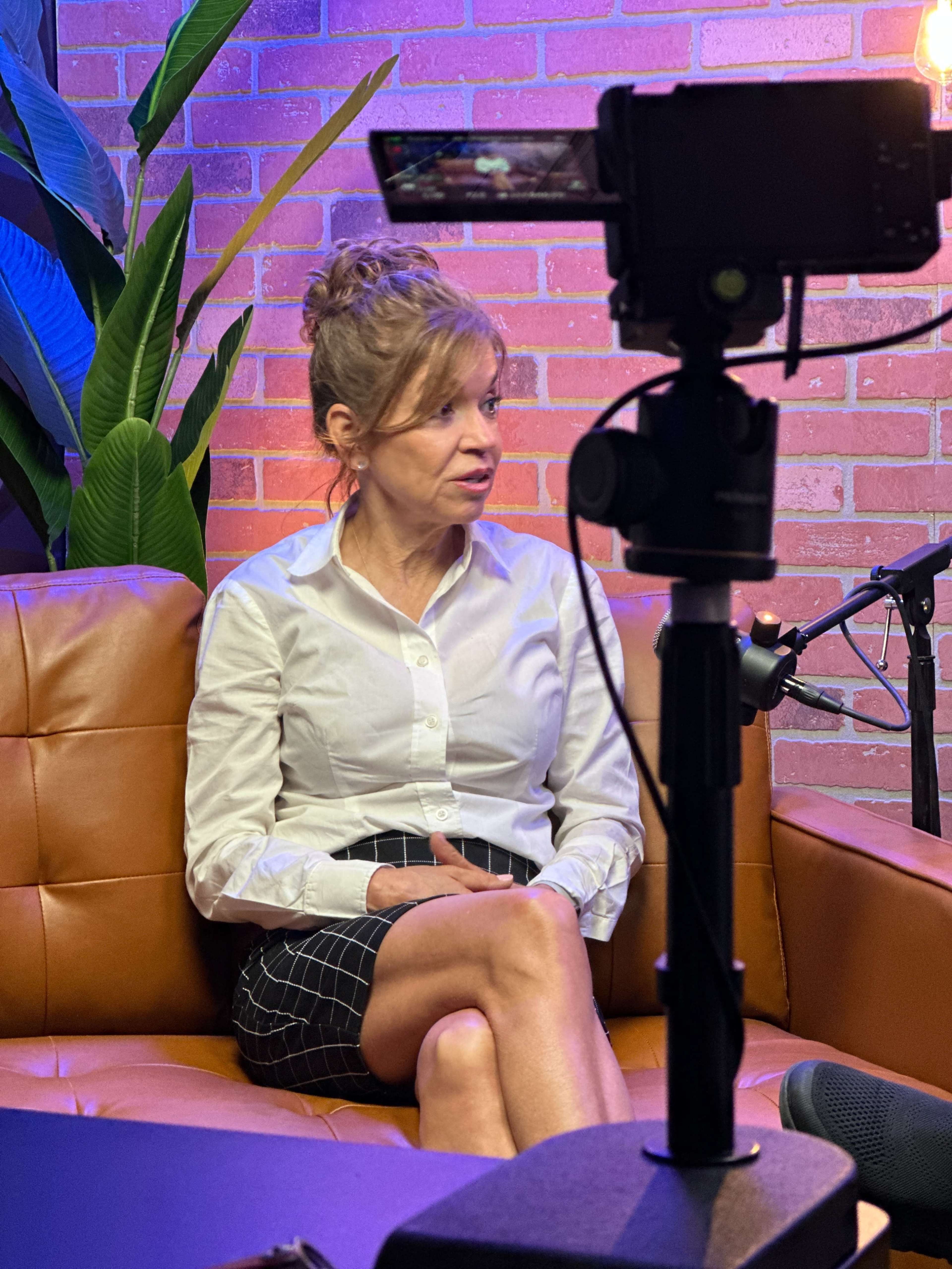 A woman in a white shirt and checkered skirt sits on a brown leather couch, with plants and a camera setup visible in the background.