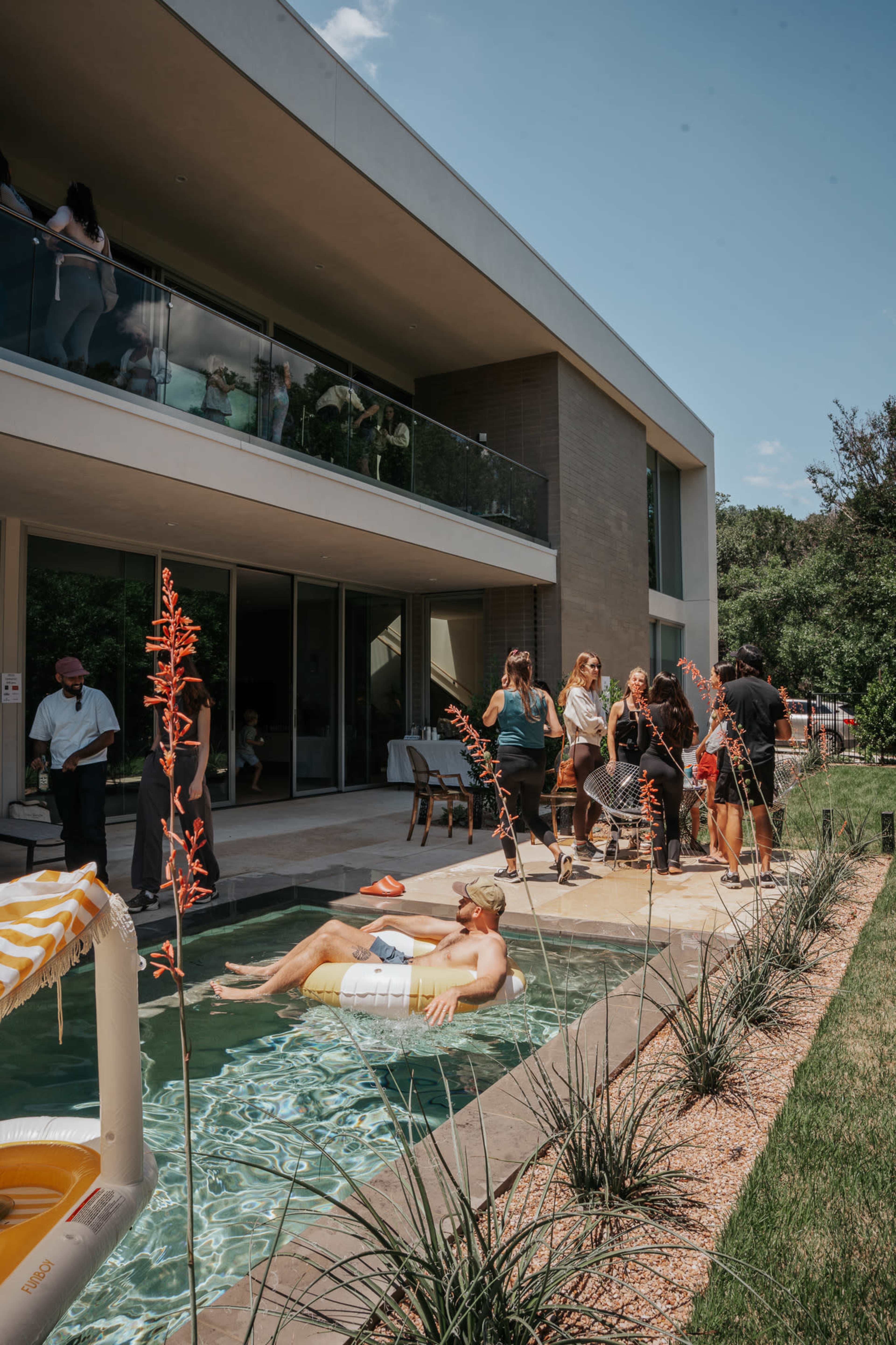 A group of people gathers around a modern house's pool area while one person relaxes in the water.