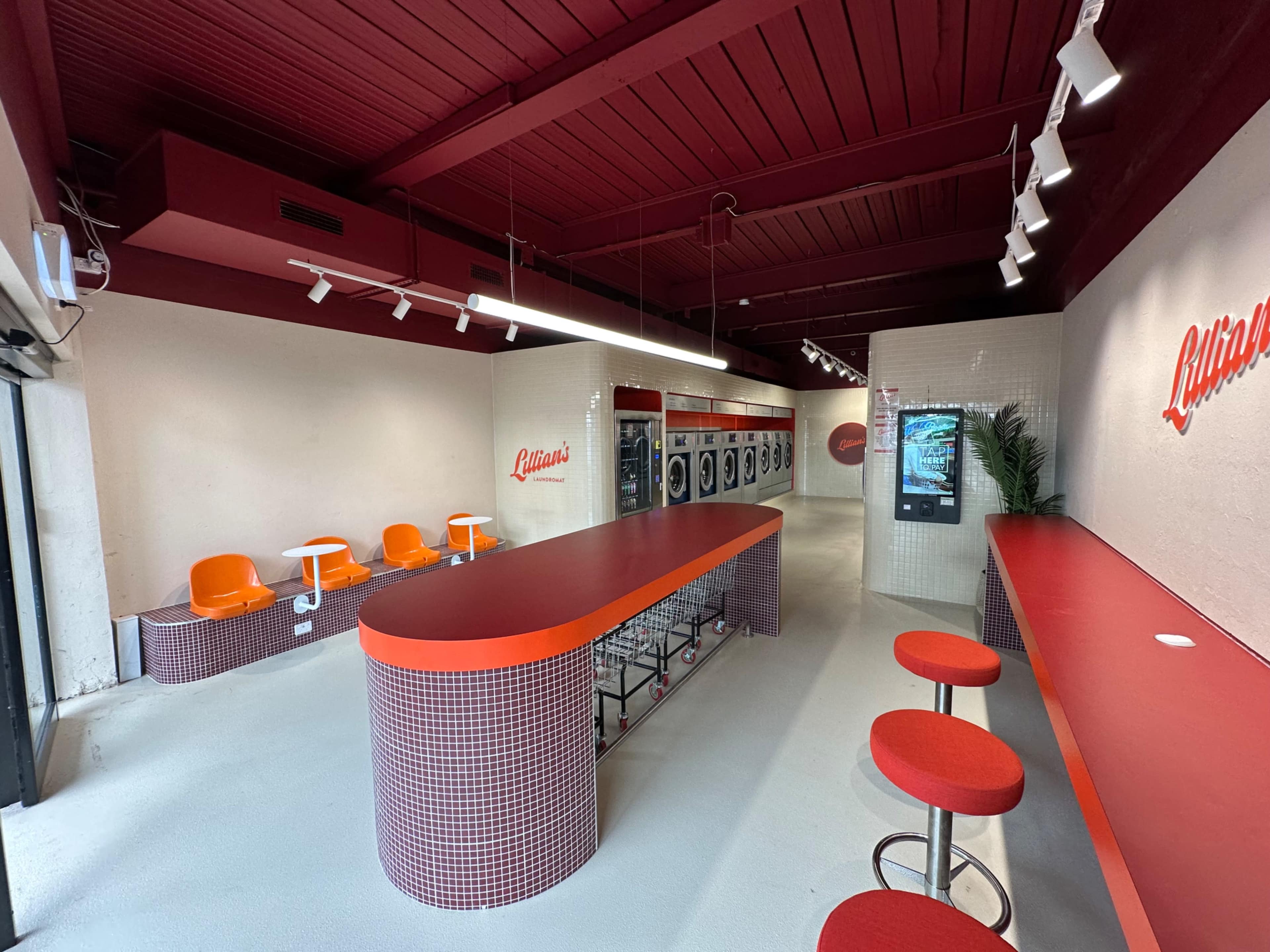 The image shows a modern laundromat with red accents, featuring a row of washing machines, seating areas, and a long countertop.