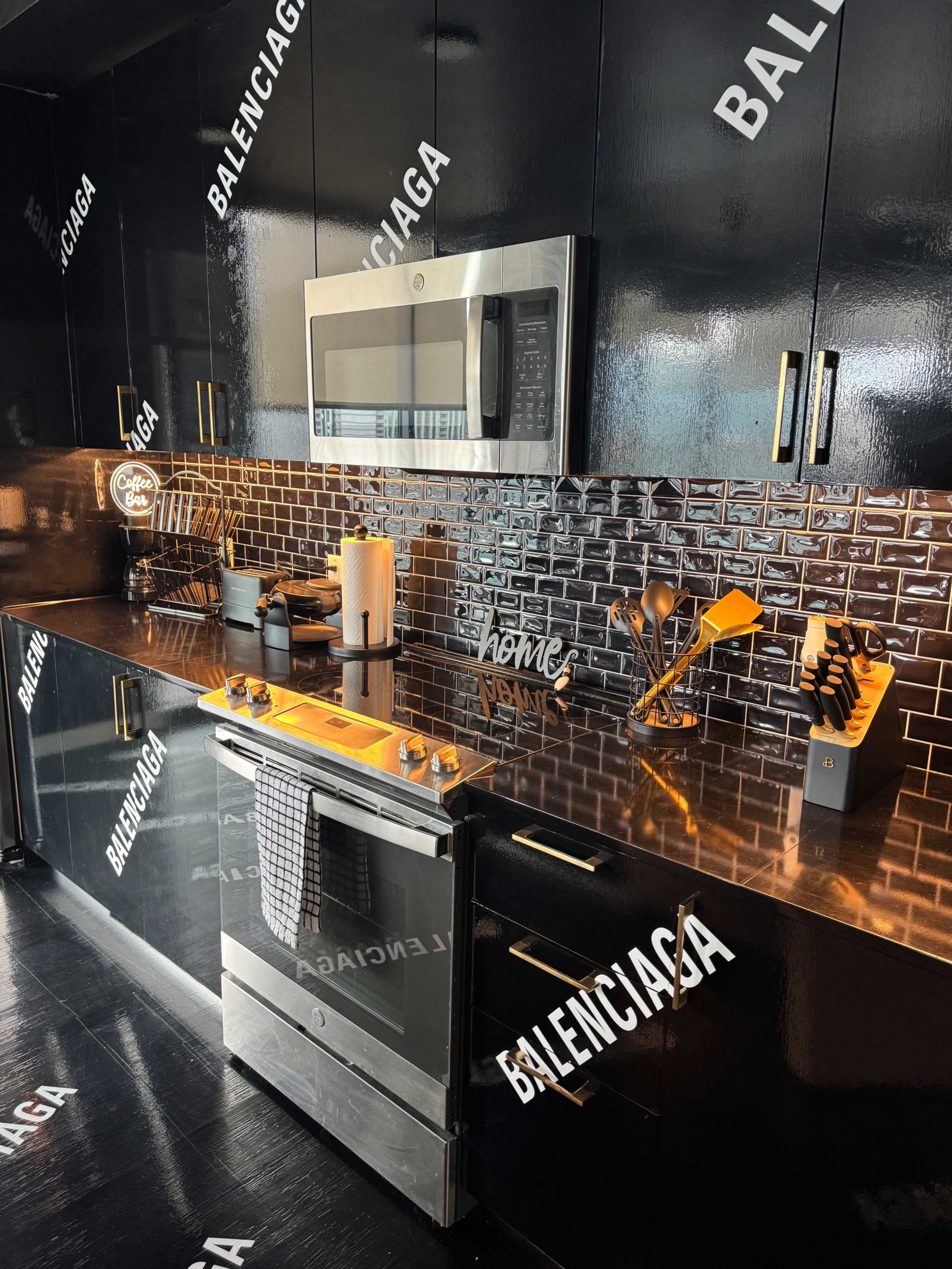 The image shows a modern kitchen with black cabinetry, a stainless steel microwave, and a stove under a shiny backsplash, all adorned with prominent "BALENCIAGA" logos.