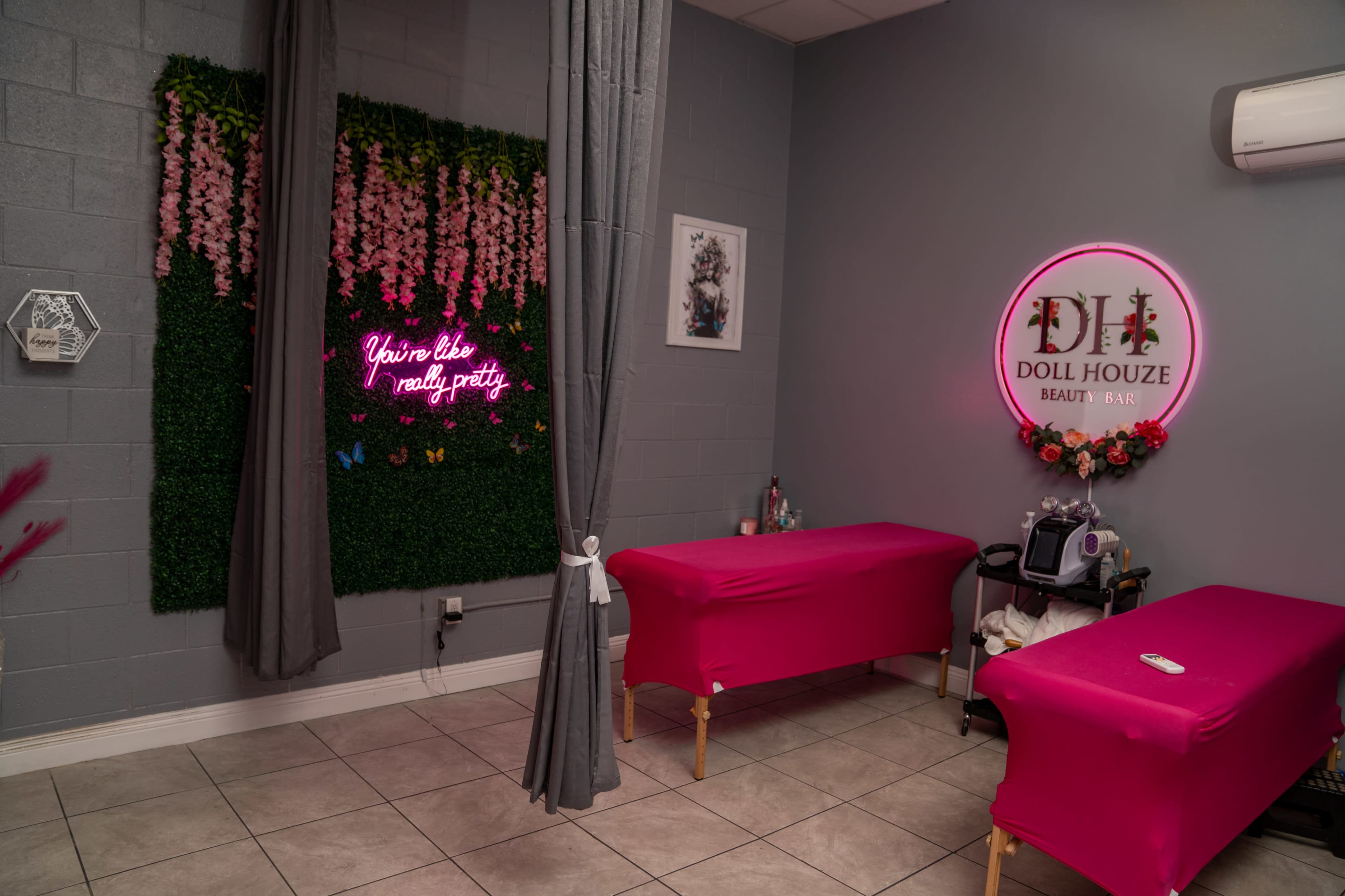 A beauty treatment room with two pink-covered massage tables, a neon sign on the wall, and decorative greenery with pink flowers.