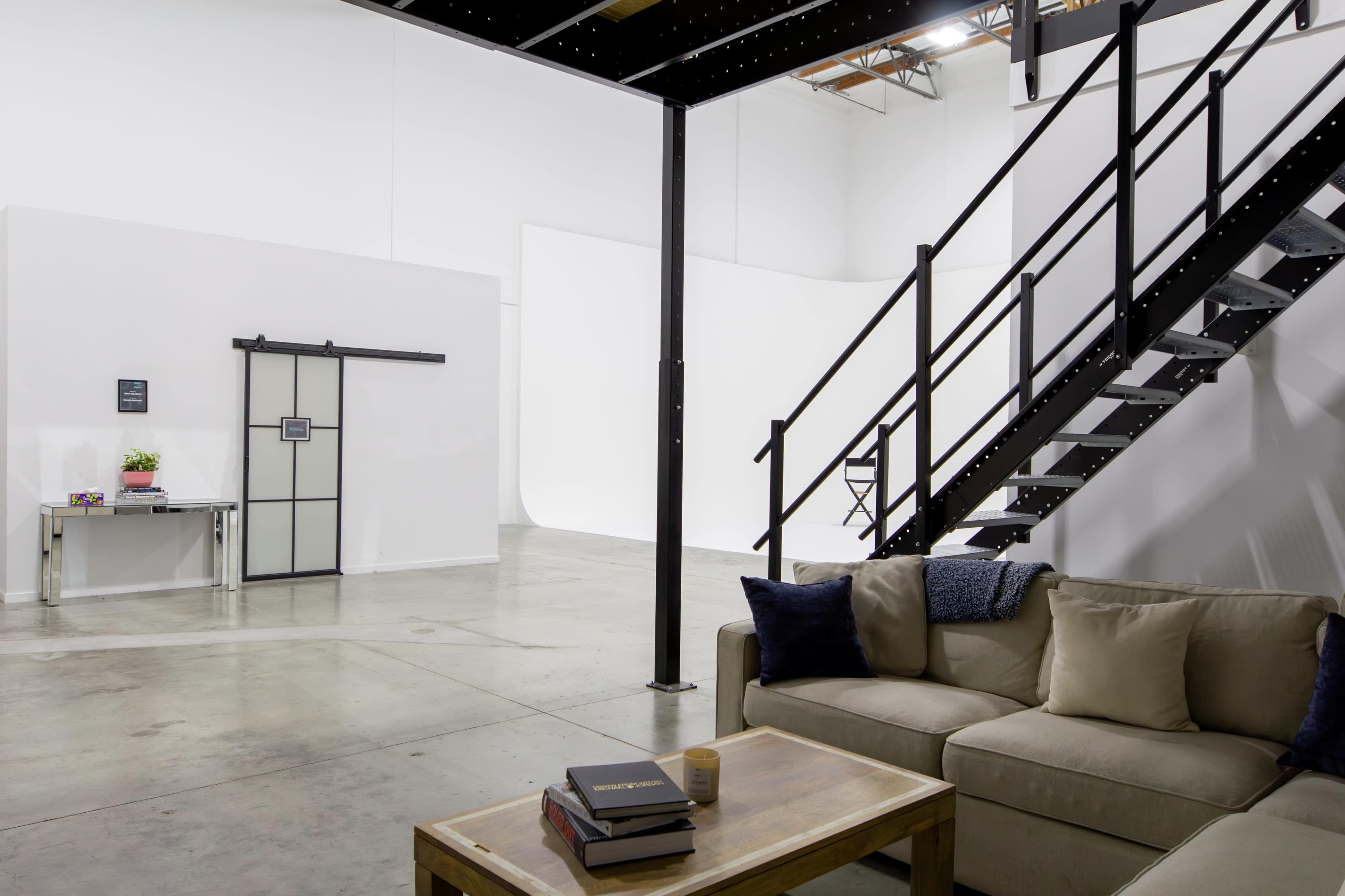 The image shows a modern, spacious interior with a seating area, a table, and a staircase leading to an upper level.