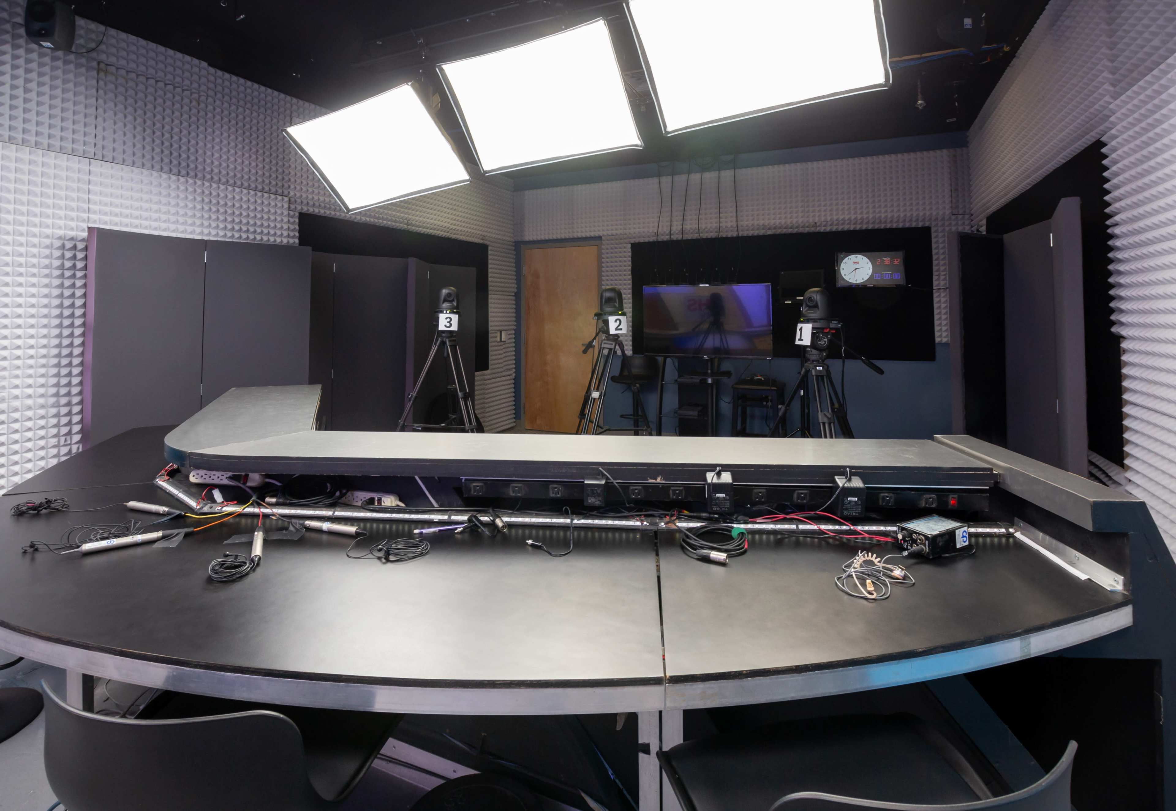 A television studio with a black table, multiple cameras, and bright overhead lighting, surrounded by soundproof wall panels.