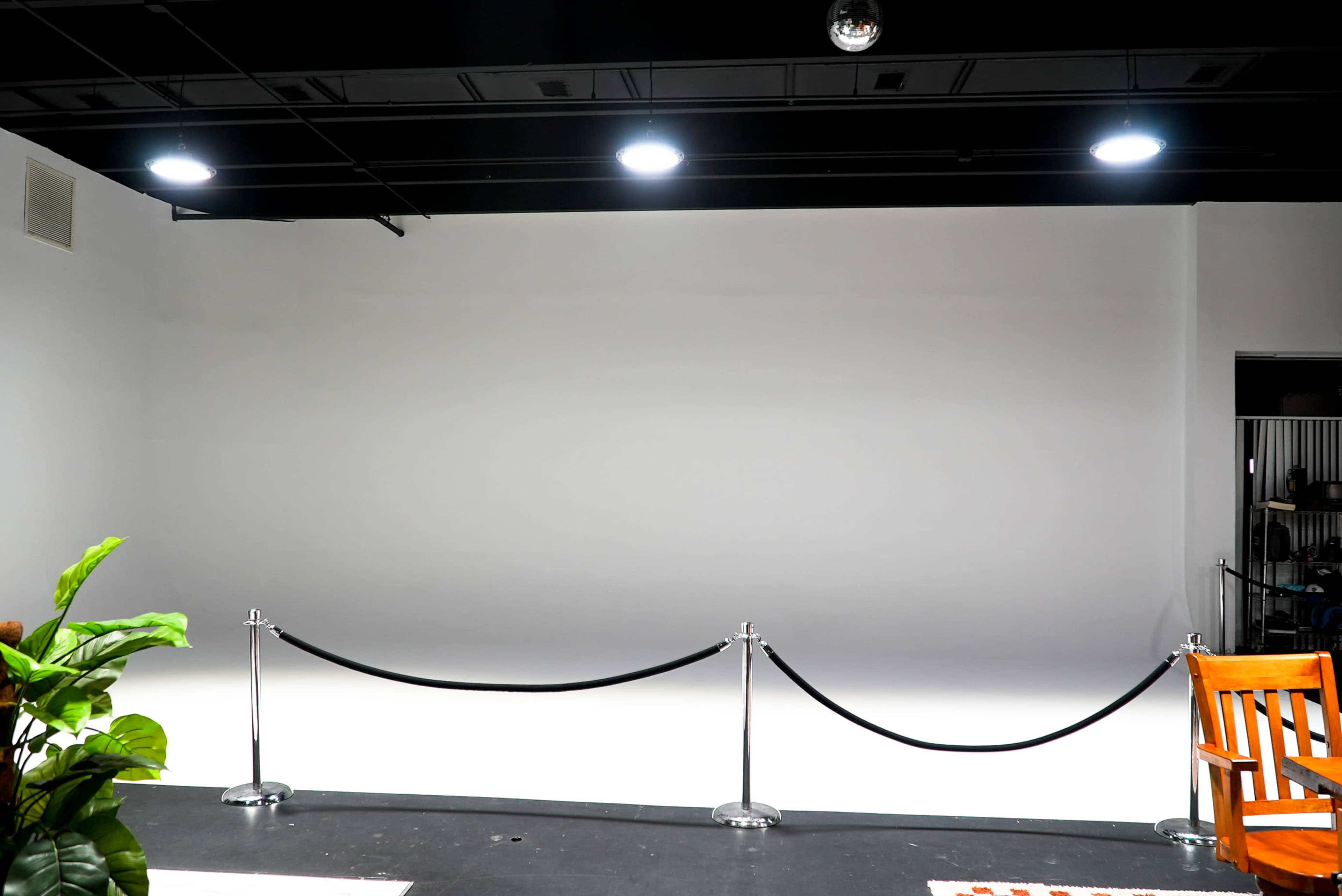 The image shows a blank studio space with a white wall, a black rope barrier, and a wooden chair in the foreground.