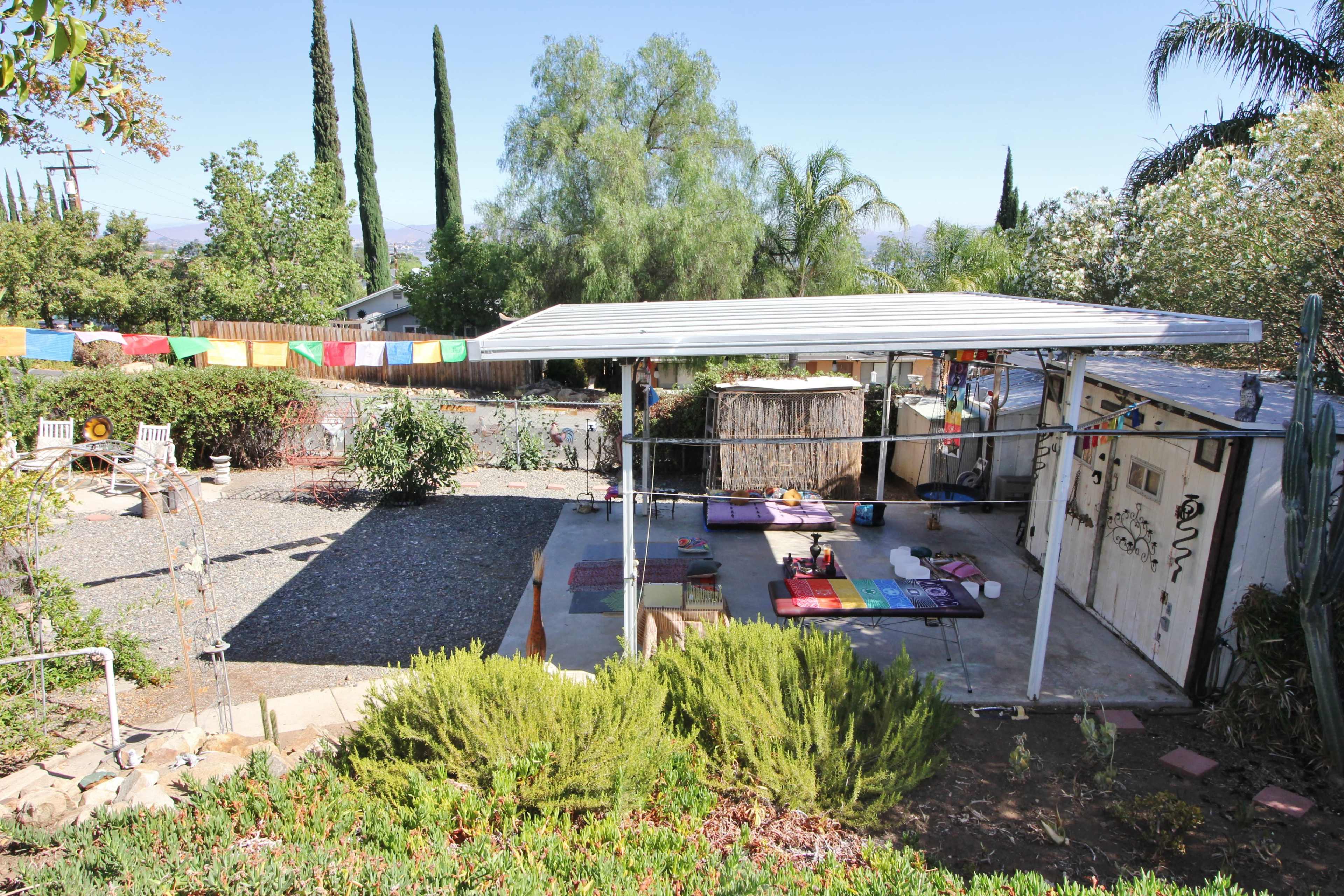 The image shows a backyard with a covered patio area, several outdoor seating options, a gravel section, and colorful prayer flags hung in the background.
