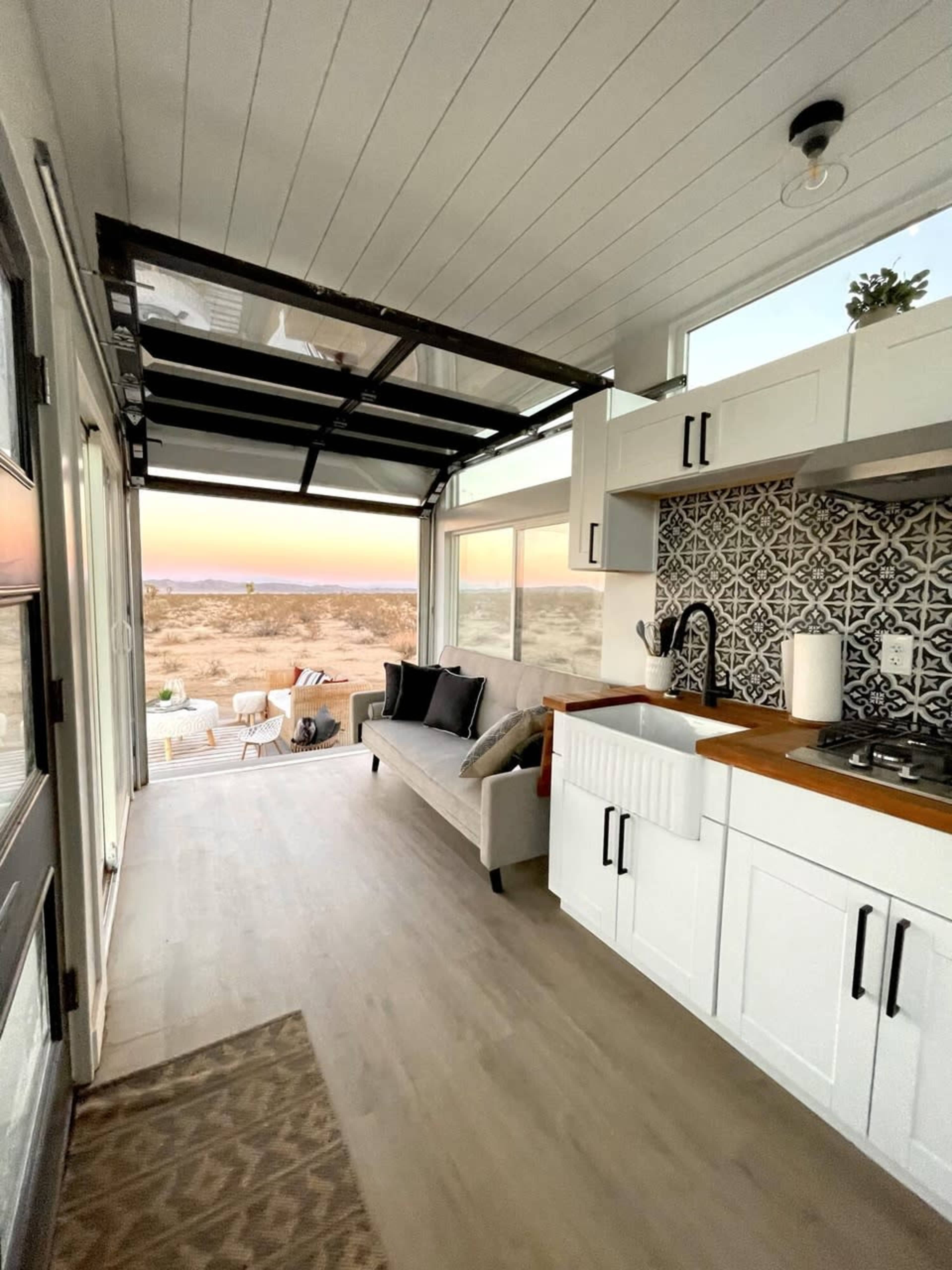 A modern interior of a tiny house with a sofa, kitchen area, and large windows overlooking a desert landscape at sunset.