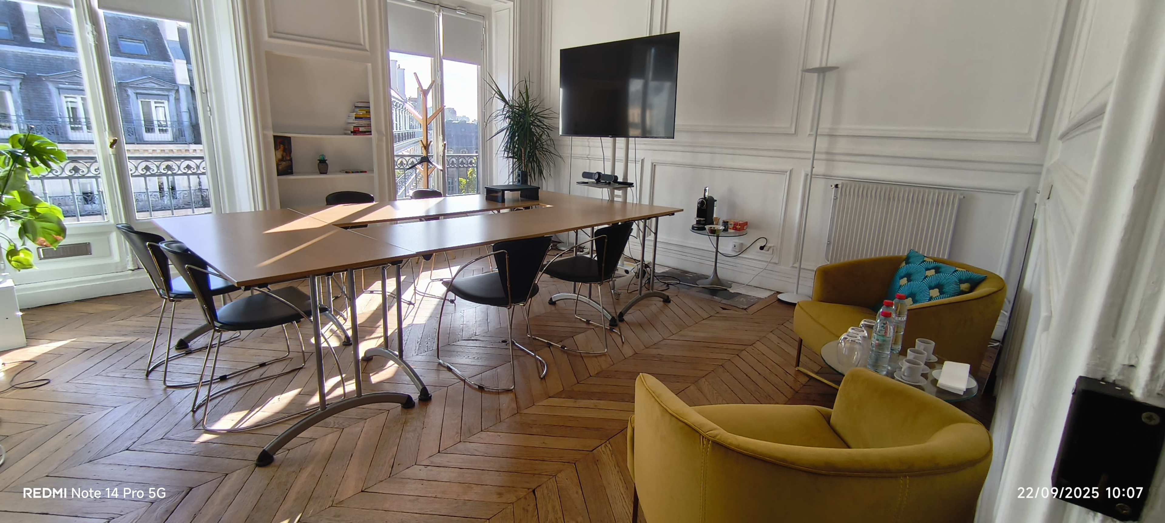 The image shows an office space with a large wooden table surrounded by black chairs, a yellow armchair in the corner, and a flat-screen TV on the wall.