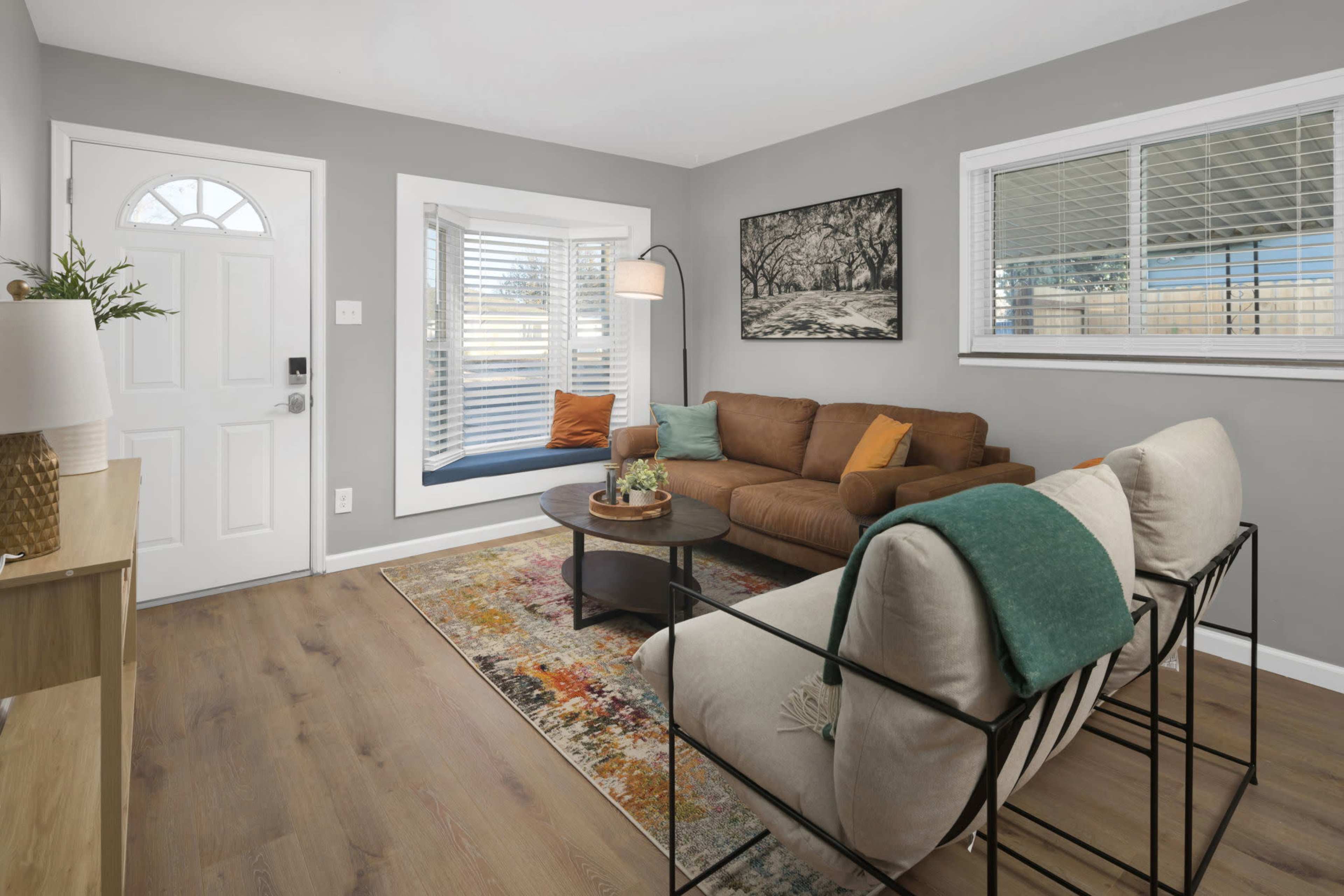 The image shows a cozy living room featuring a brown sectional sofa, a round black coffee table, and a decorative rug on wooden flooring, with a large window providing natural light.