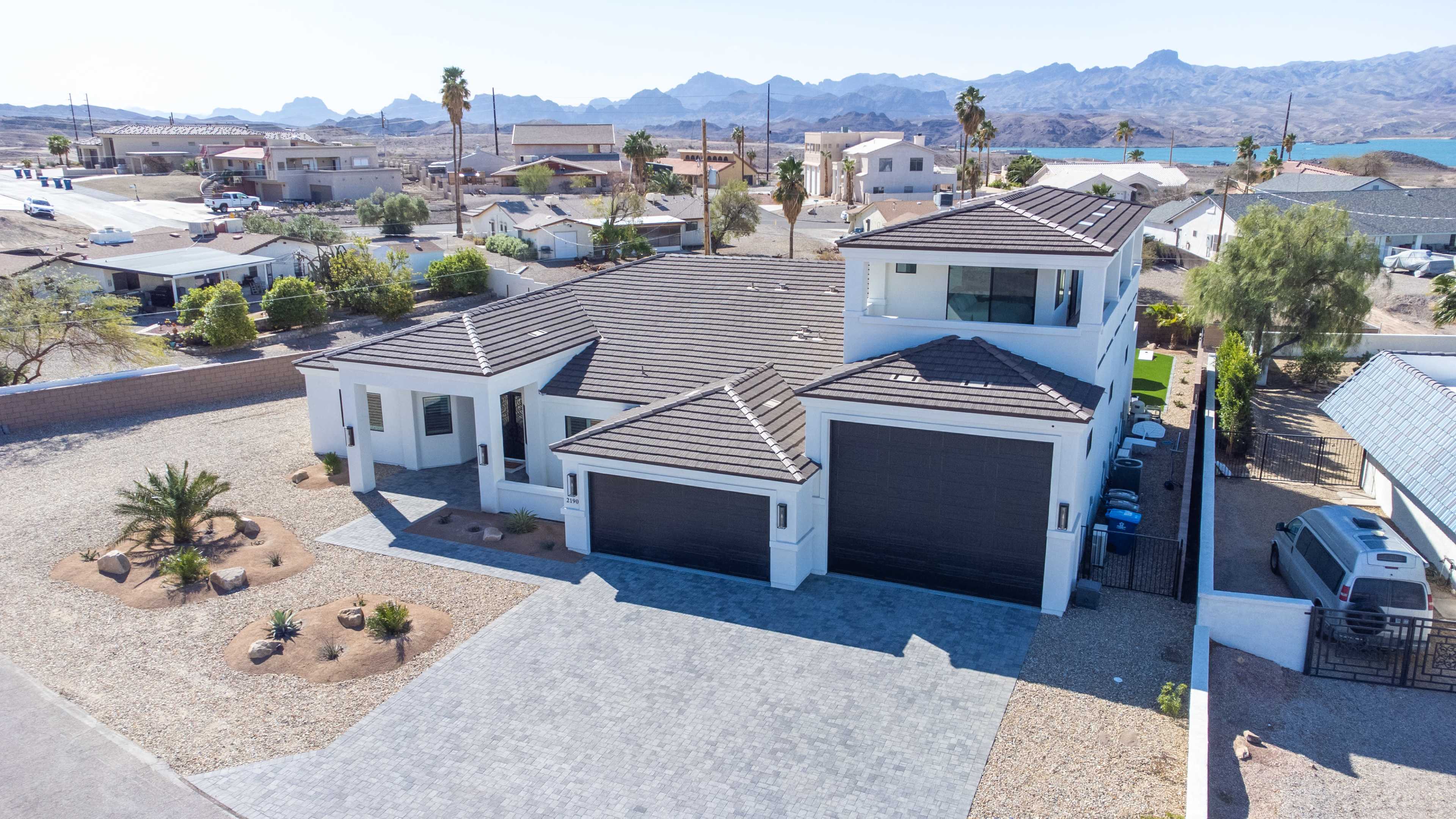 A modern white house with a dark tiled roof and a landscaped yard, situated in a desert area with mountains in the background.