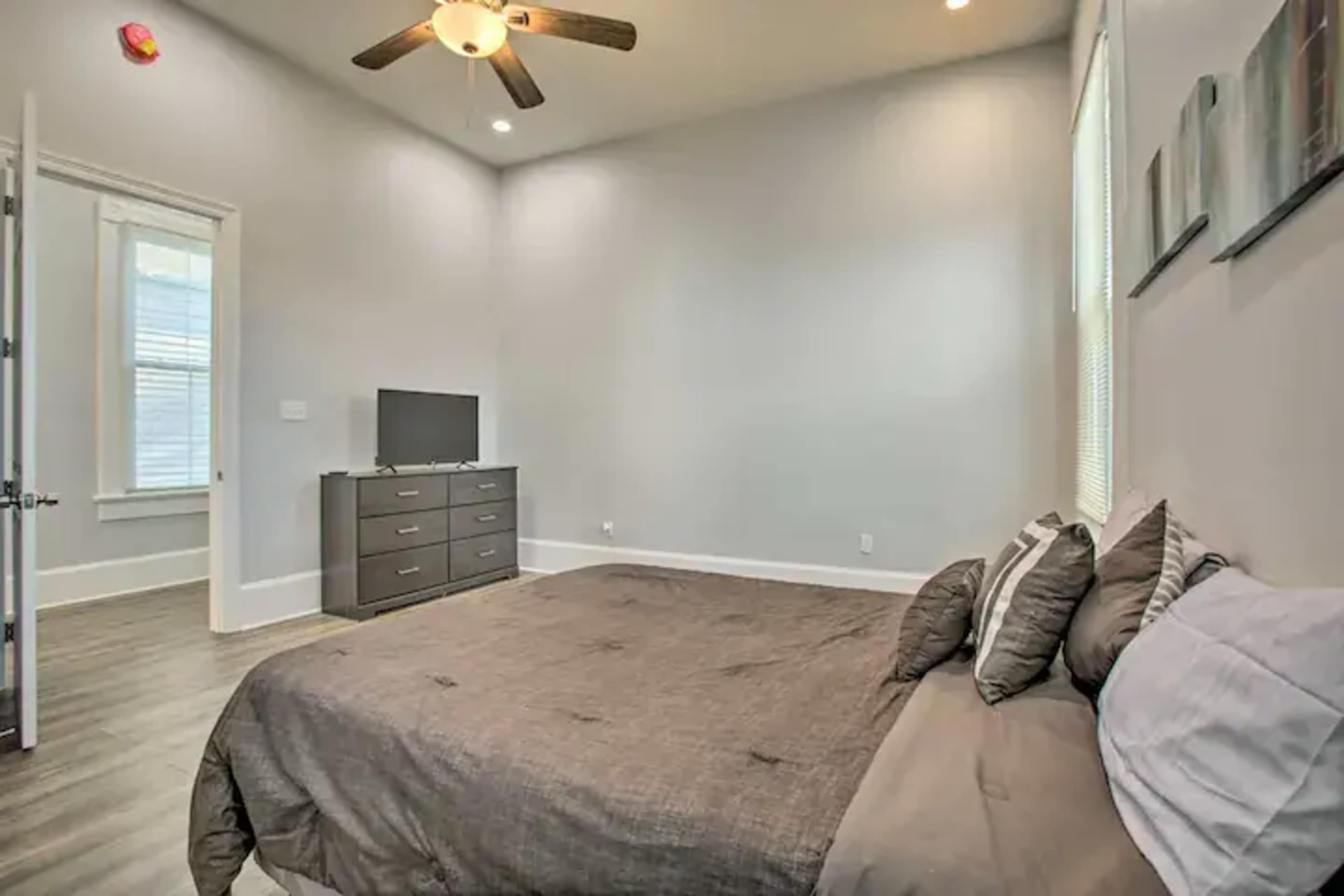 A tidy bedroom with a gray bedspread, a dresser, a television, and natural light coming through the windows.