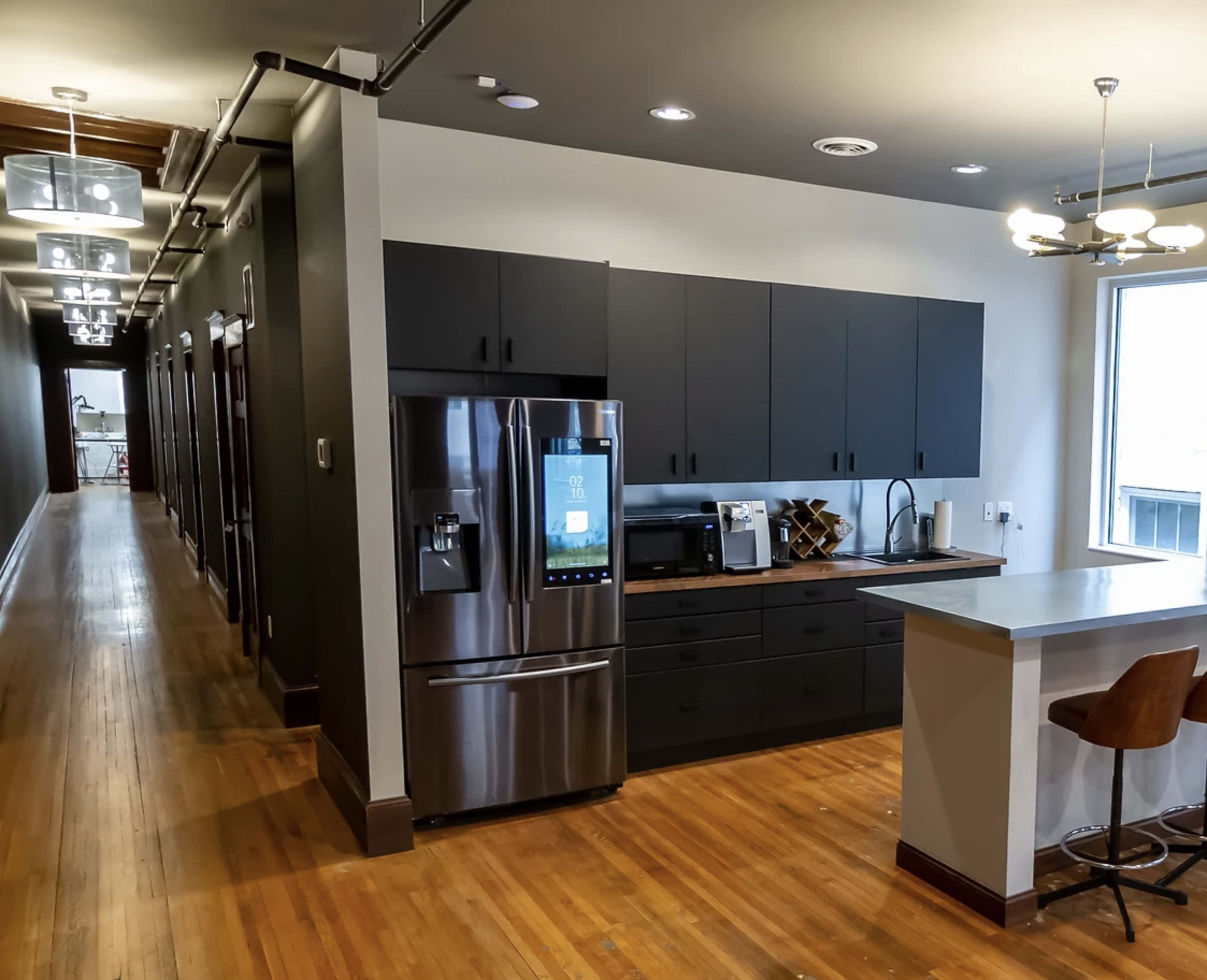 A modern kitchen with dark cabinetry, a stainless steel refrigerator, and a bar counter, leading to a hallway with several doors.