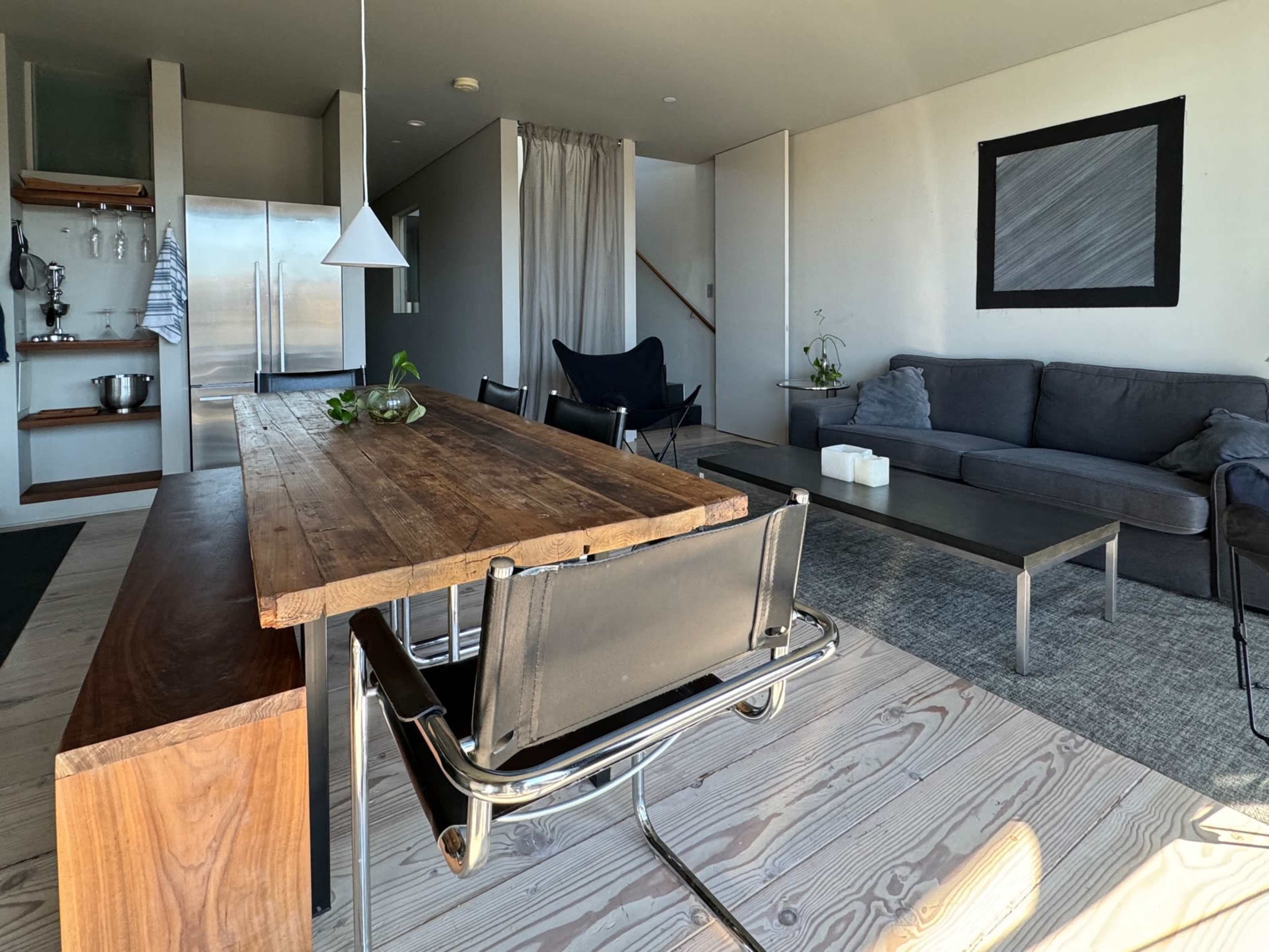 A modern interior with a wooden dining table, black chairs, a gray sofa, a coffee table, and a kitchenette in a spacious, well-lit room.