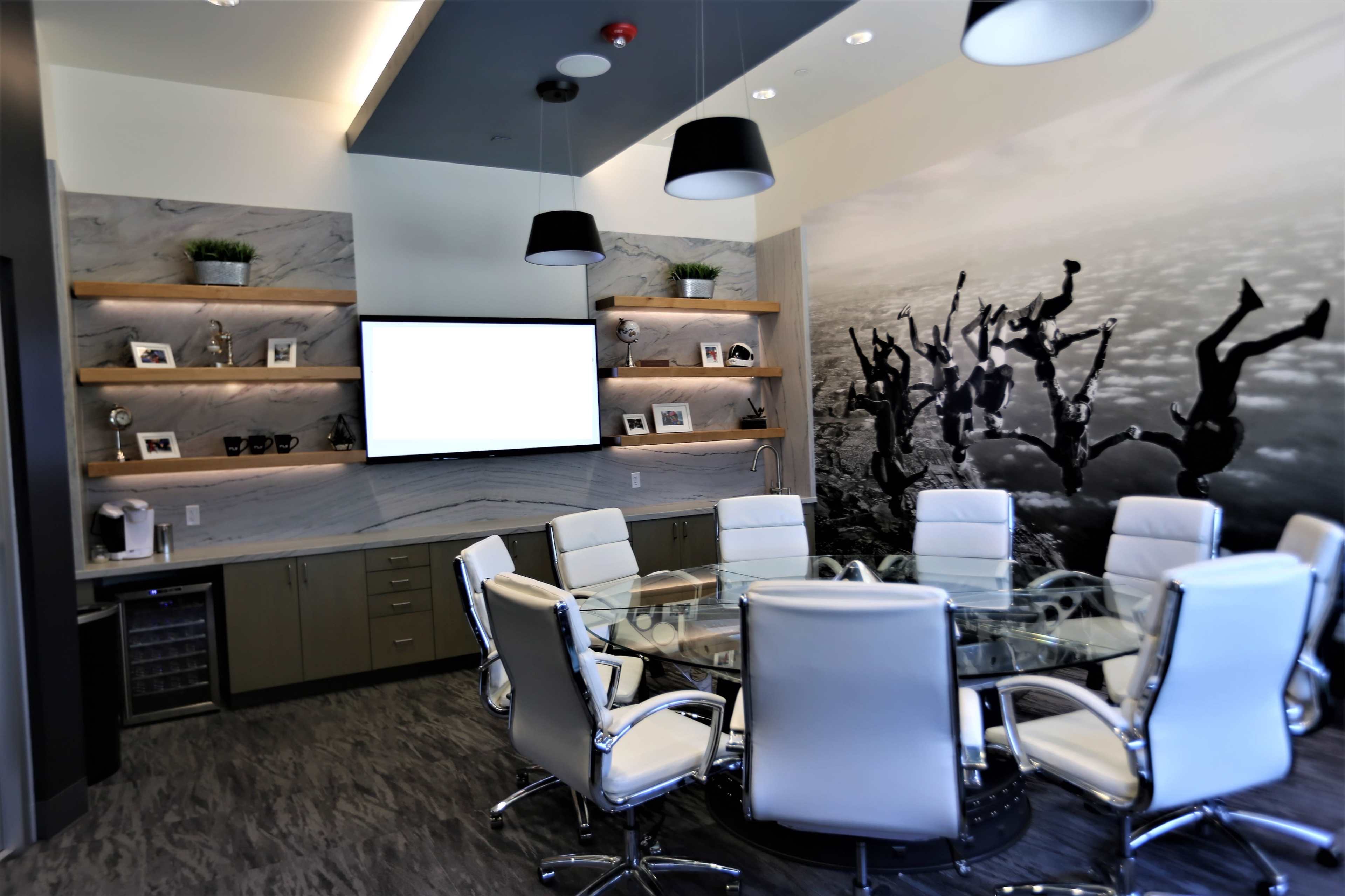The image shows a modern conference room with a circular glass table, white chairs, and shelves displaying framed photos, alongside a large wall mural of people jumping into water.