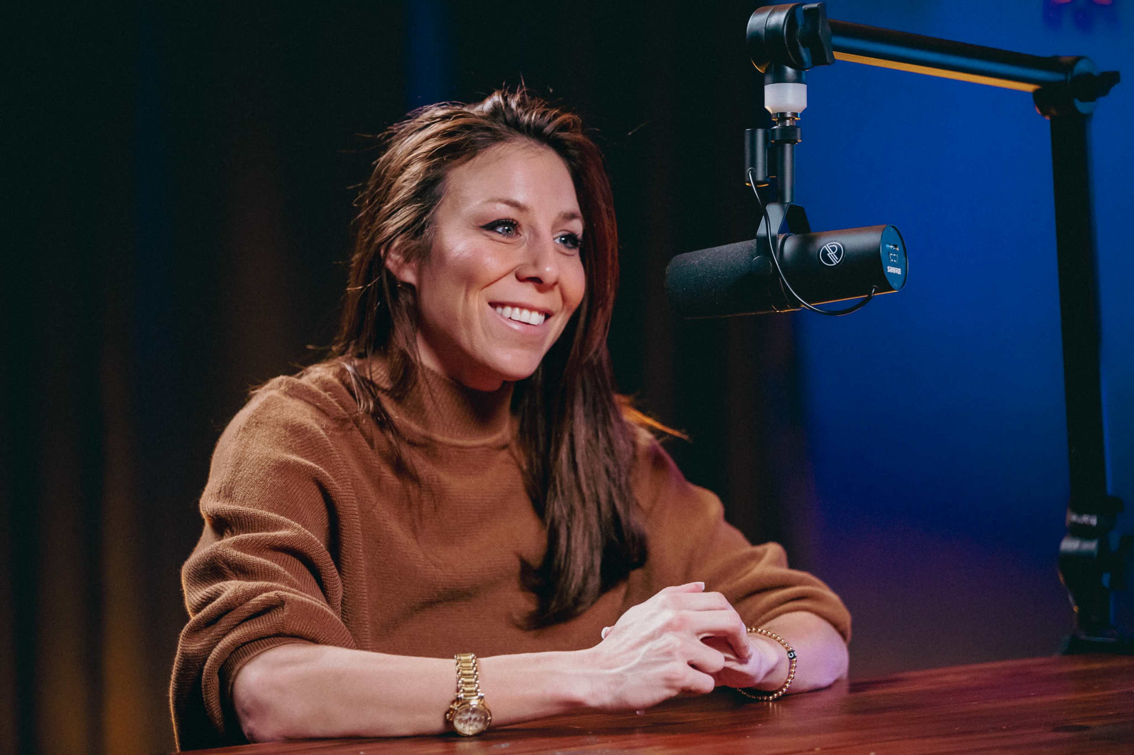 A woman with long brown hair sits at a table with a microphone in front of her, smiling.