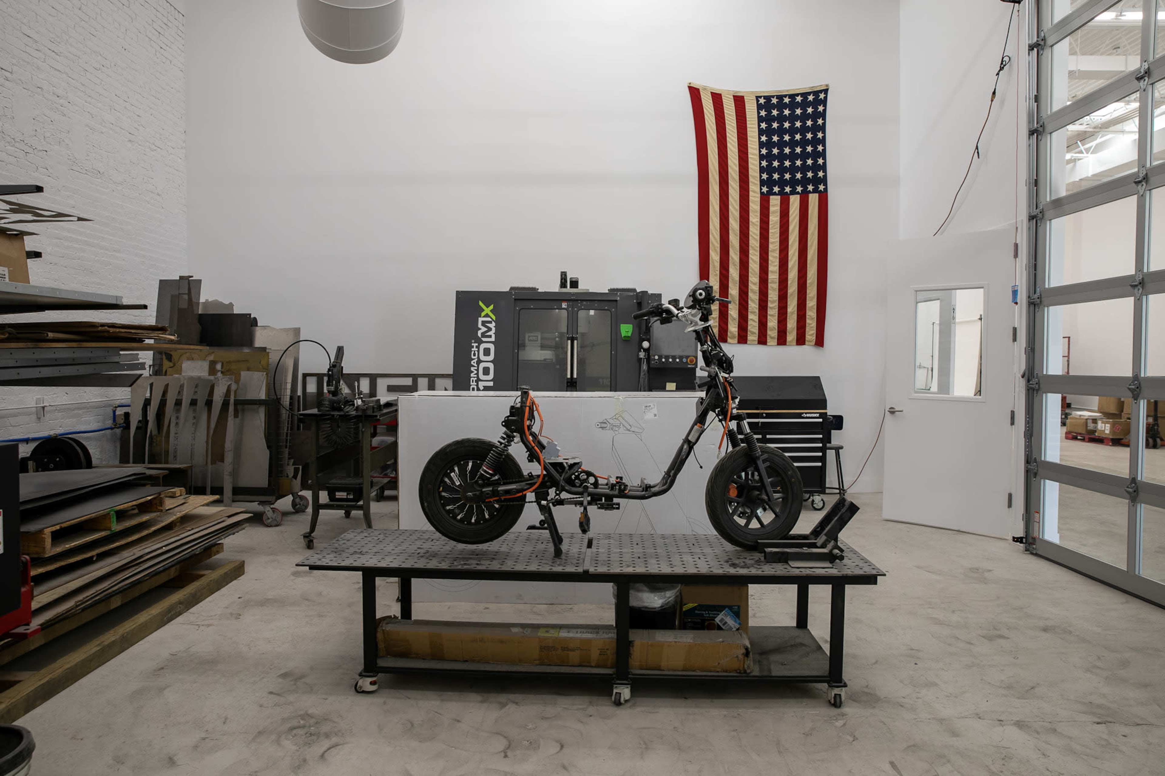 An electric scooter is prominently displayed on a metal work table in a well-lit workshop, with an American flag hanging on the wall behind it.