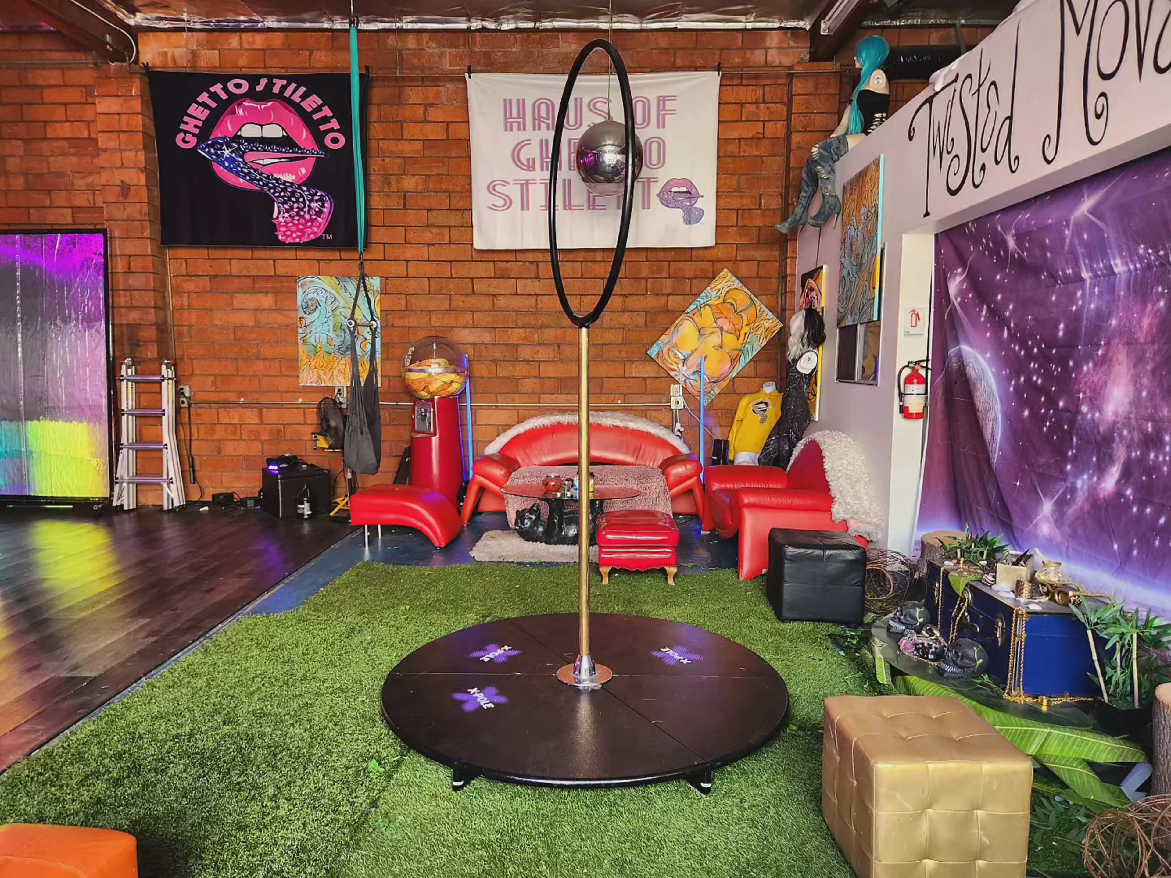The image shows a studio space featuring a pole in the center, surrounded by colorful decor, plush seating, and artistic murals on the walls.