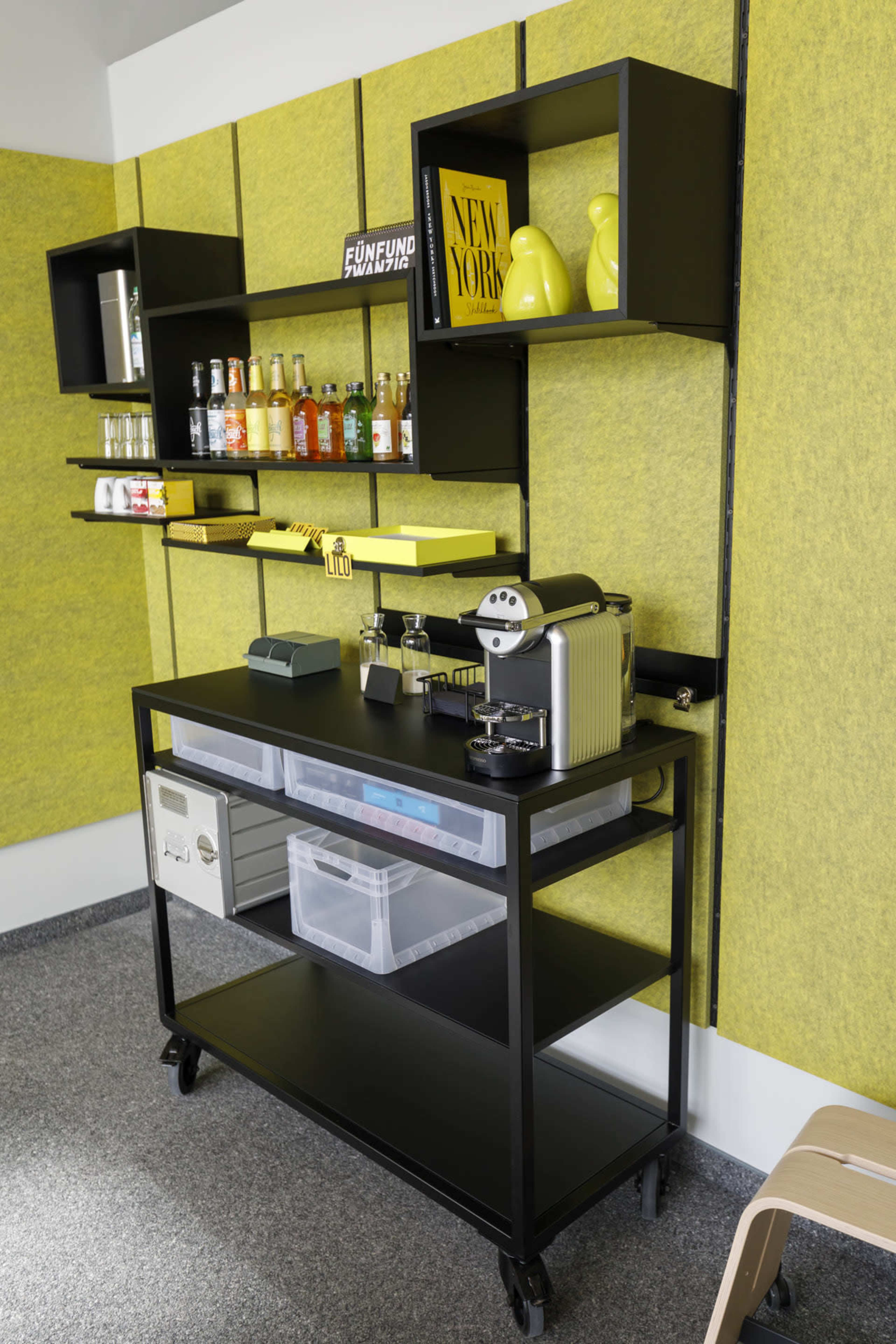 The image shows a black mobile cart with a clear storage bin on the bottom, topped with a beverage display mounted on a wall that features yellow accents.