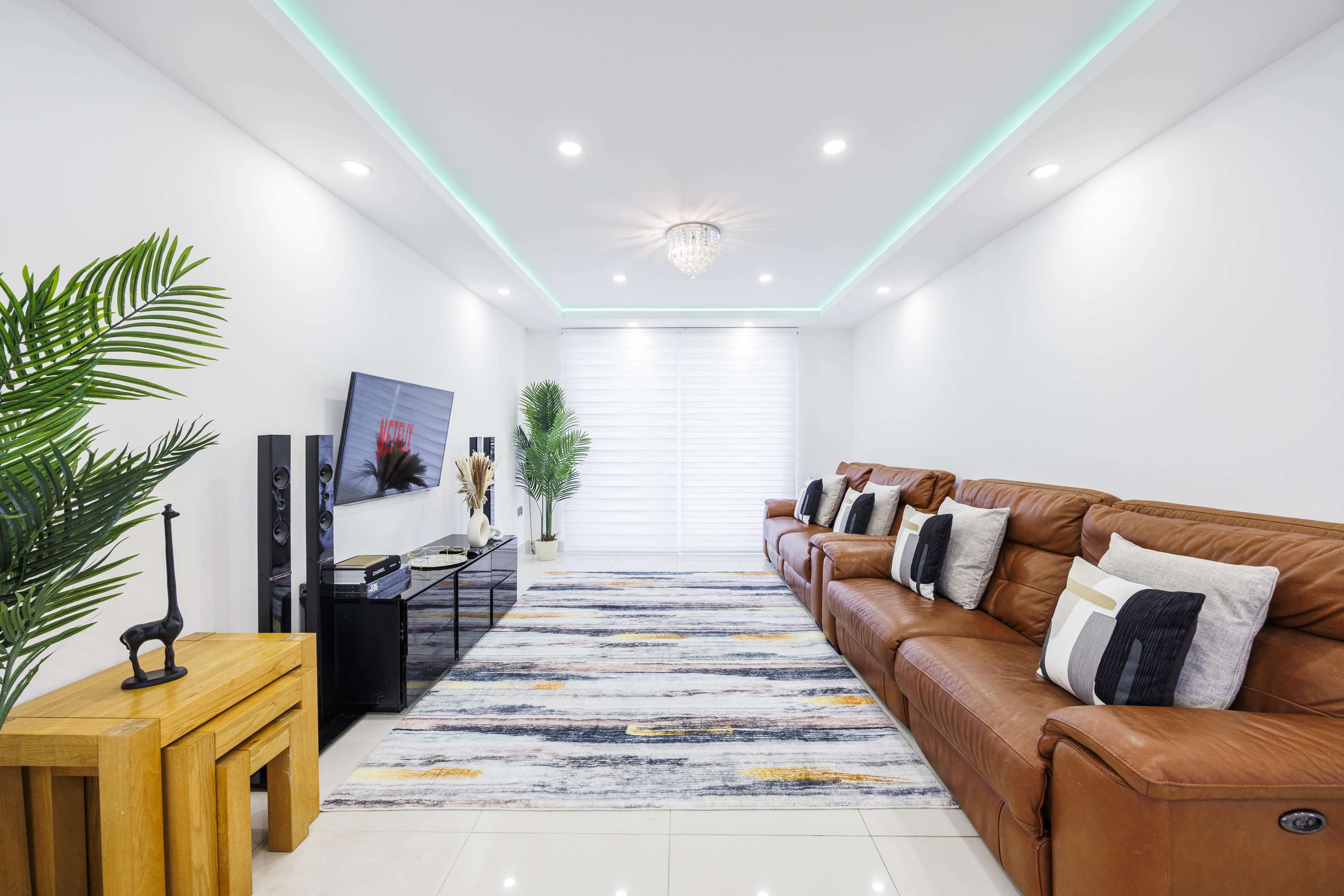 A modern living room features a brown leather sofa, a wooden side table, a large area rug, and a television on a sleek entertainment unit, illuminated by recessed lighting and a crystal chandelier.